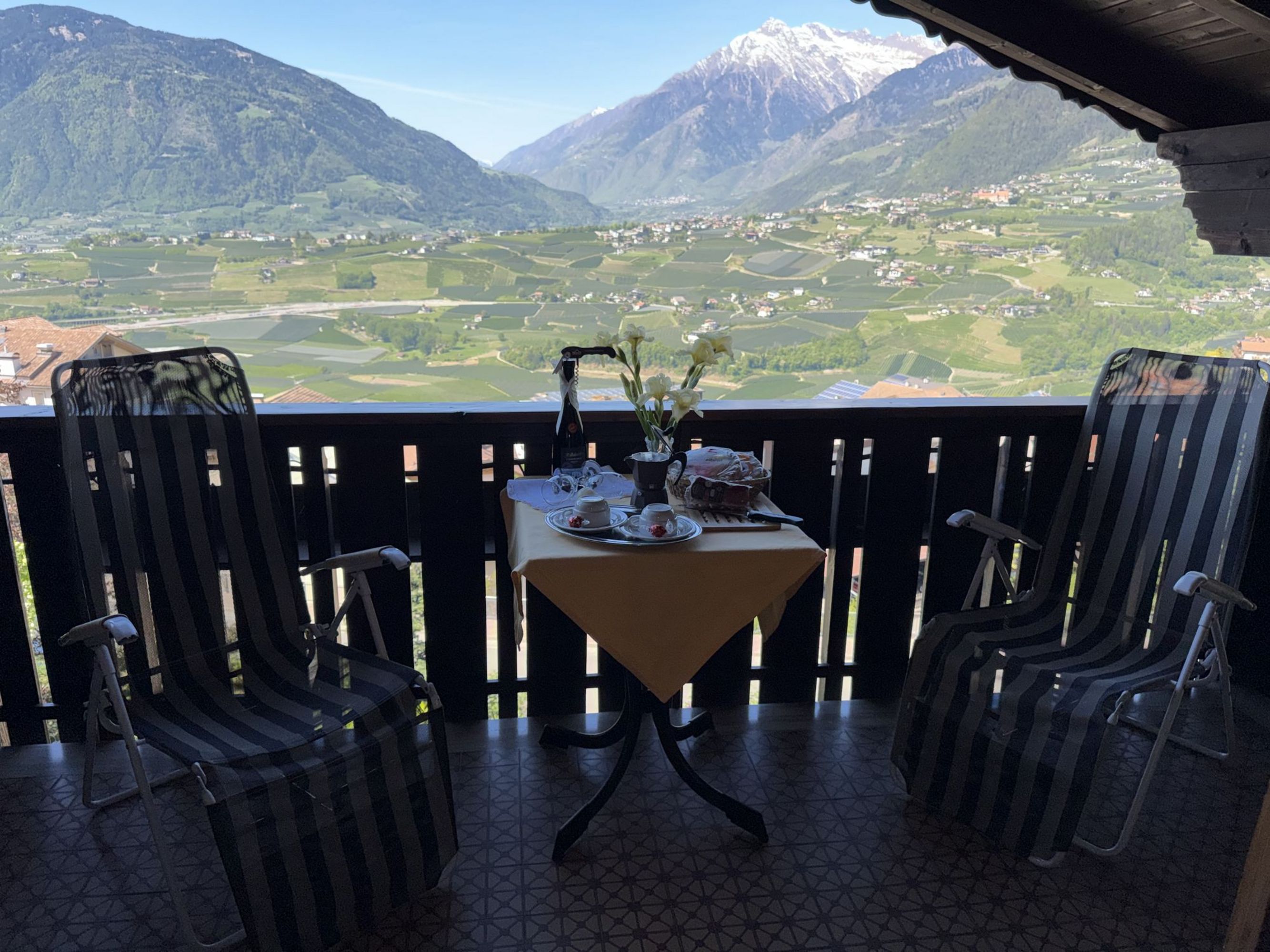 Zwei Gartenstühle stehen auf einem Balkon vor einem kleinen runden Tisch mit Kaffeegedeck und Blumen, im Hintergrund erstreckt sich ein Tal mit Bergen.