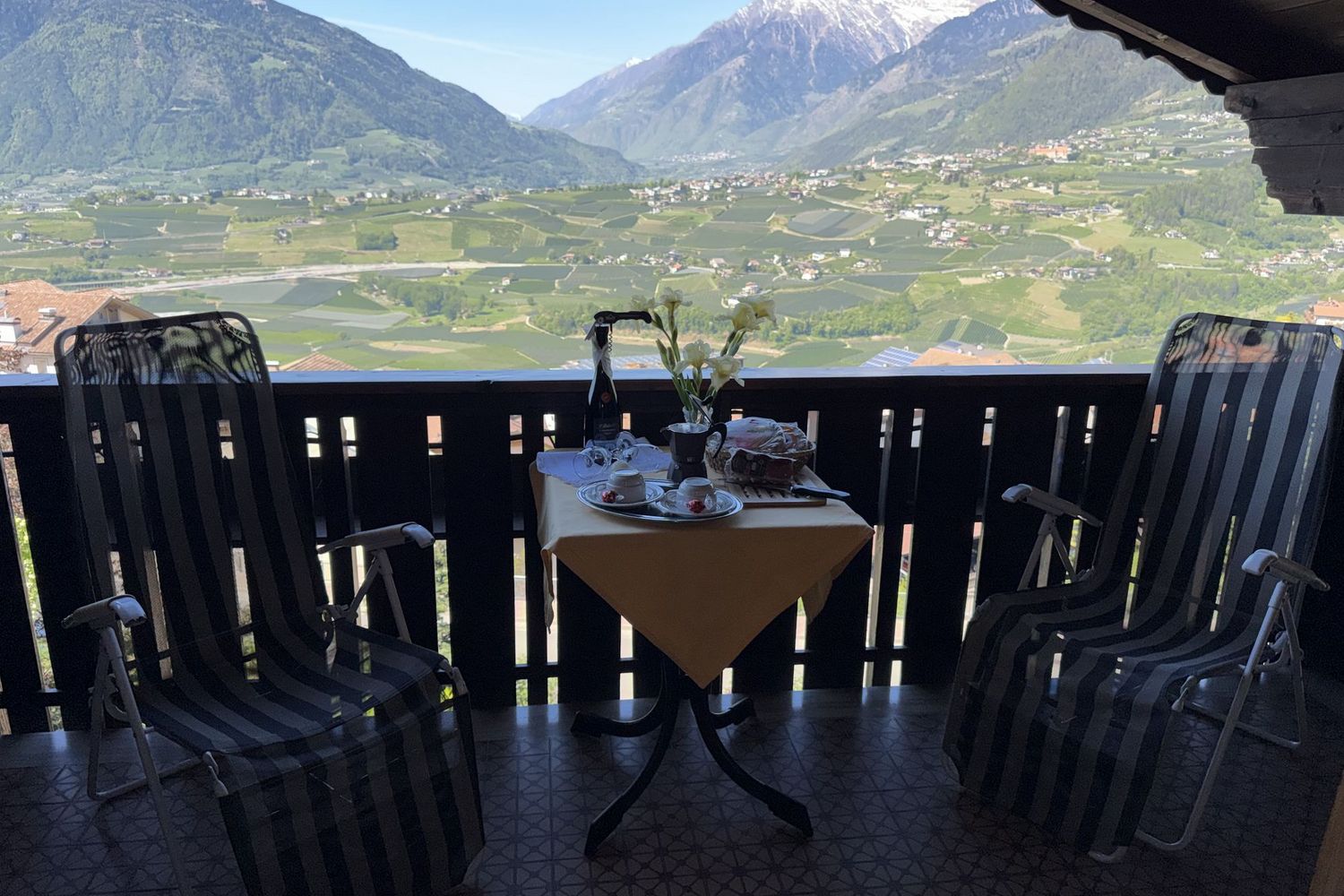 Zwei Gartenstühle stehen auf einem Balkon vor einem kleinen runden Tisch mit Kaffeegedeck und Blumen, im Hintergrund erstreckt sich ein Tal mit Bergen.