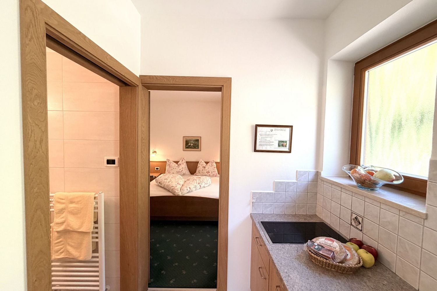 Ein Blick durch eine Tür in ein Schlafzimmer mit Doppelbett, während im Vordergrund eine kleine Kochnische mit Herdplatte und einem Obstkorb zu sehen ist.