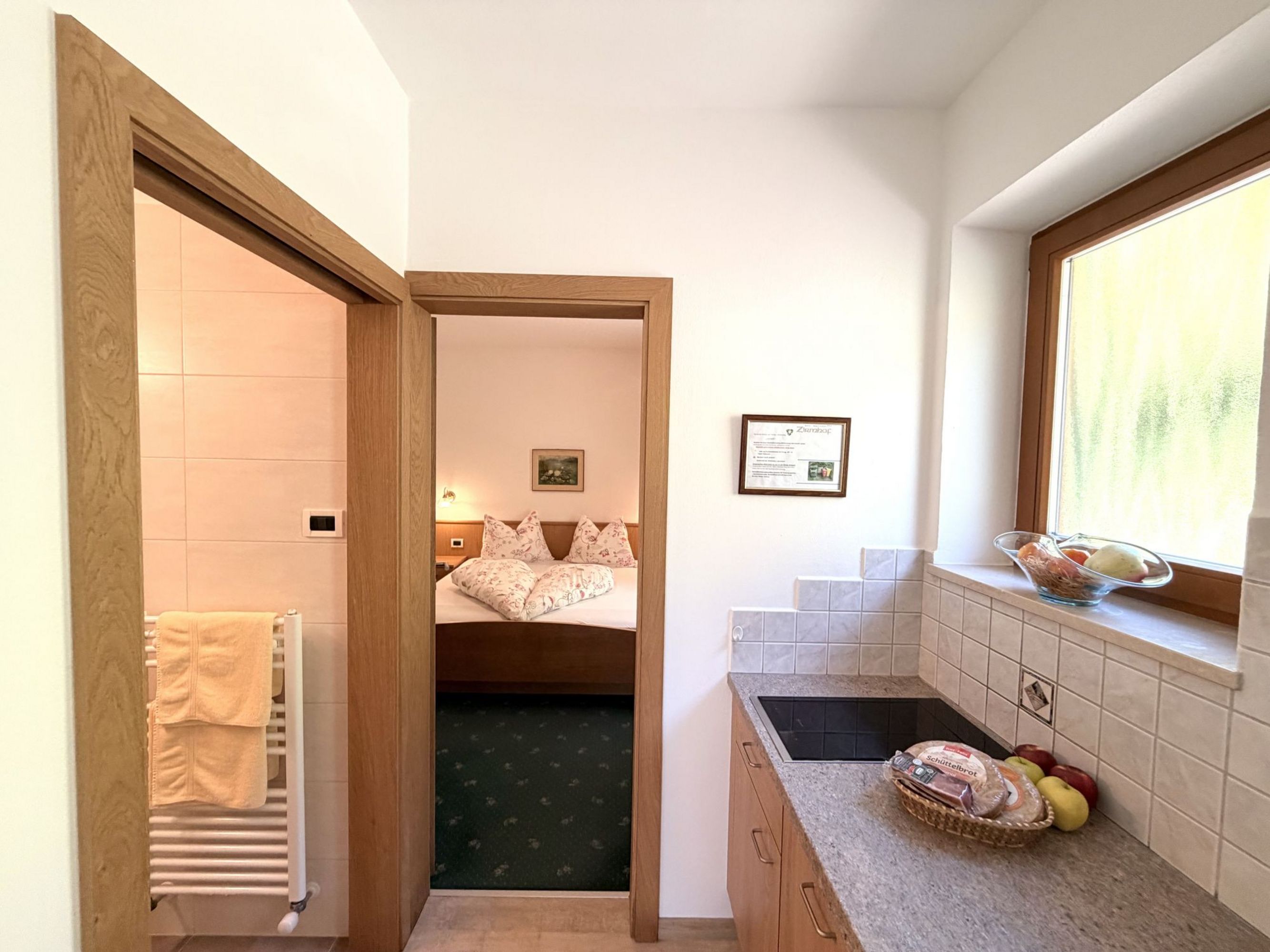 Ein Blick durch eine Tür in ein Schlafzimmer mit Doppelbett, während im Vordergrund eine kleine Kochnische mit Herdplatte und einem Obstkorb zu sehen ist.