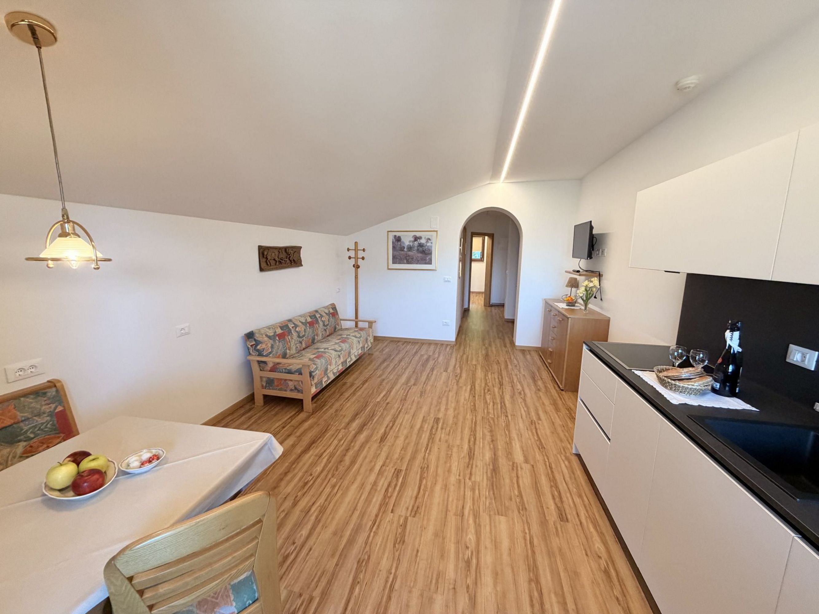An open living area including a kitchenette and dining space with a sofa under a sloping roof, light wood flooring, and a doorway to the hallway.