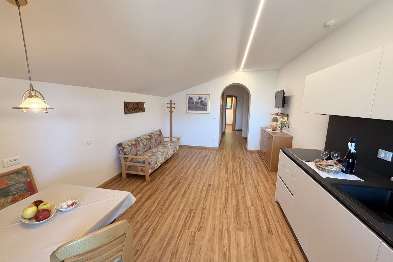 An open living area including a kitchenette and dining space with a sofa under a sloping roof, light wood flooring, and a doorway to the hallway.