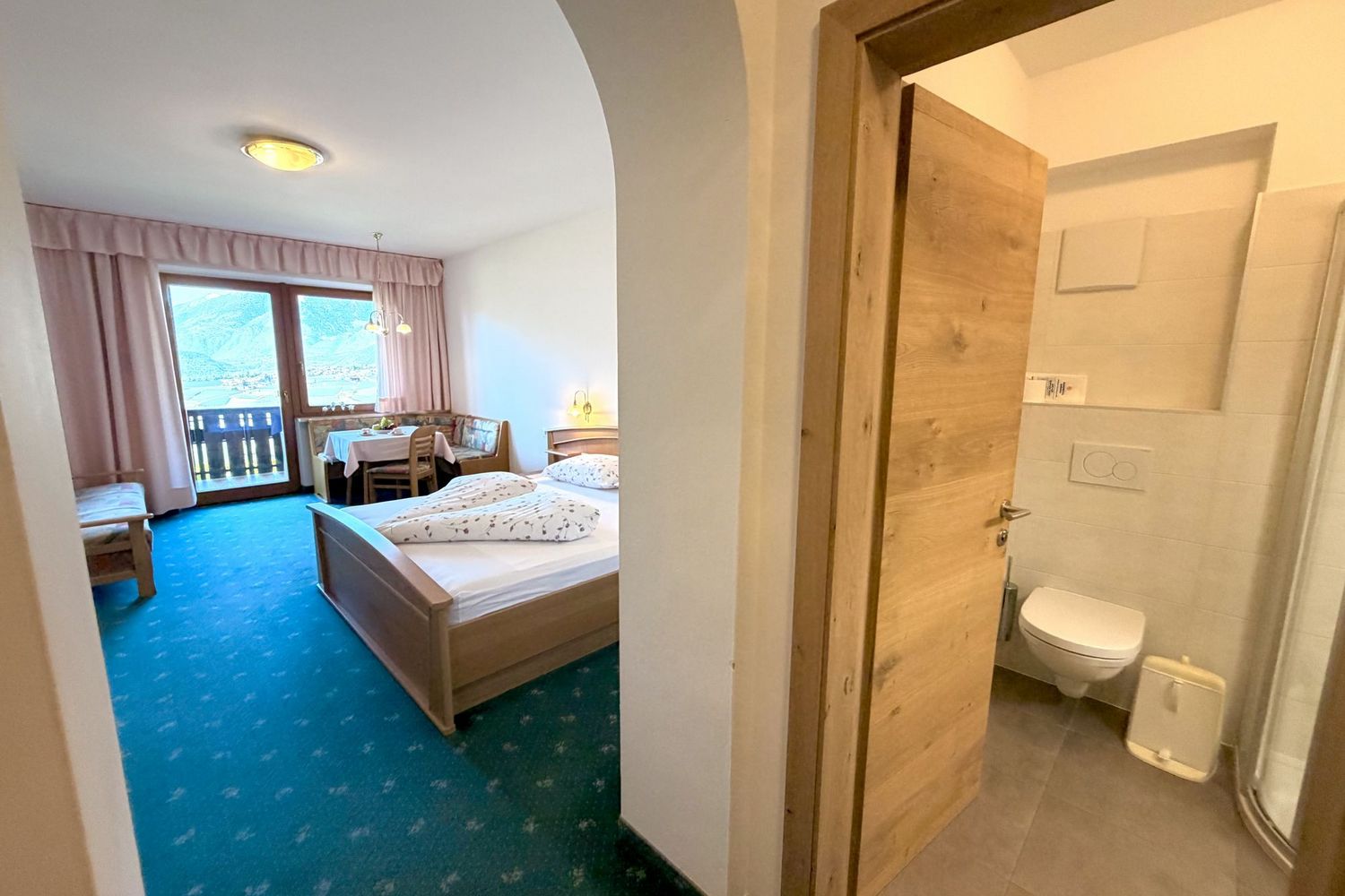 Ein Hotelzimmer mit Doppelbett, Sitzecke und einem Balkon, daneben ein gefliestes Badezimmer mit WC, gesehen durch einen Torbogen.