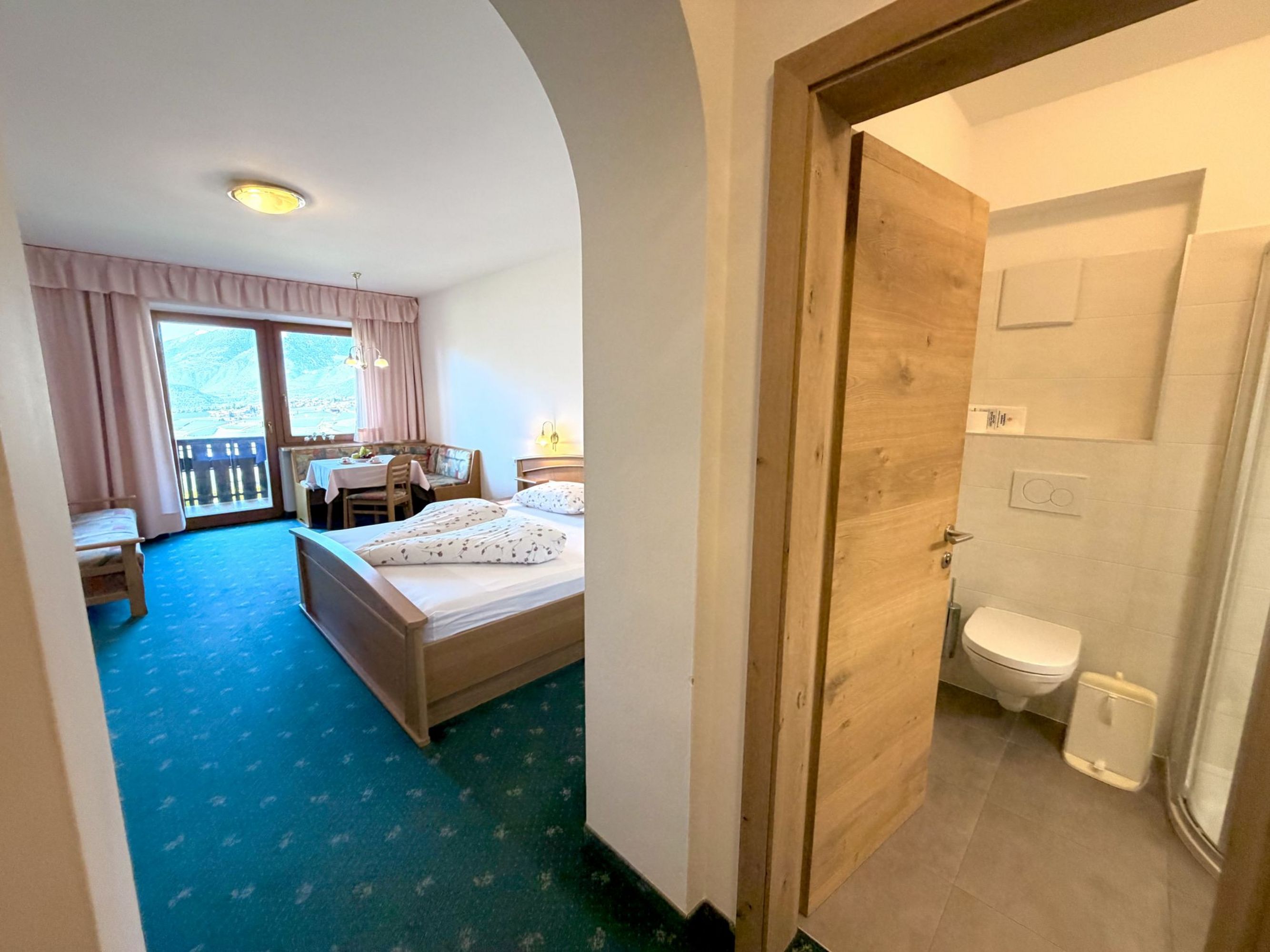 Ein Hotelzimmer mit Doppelbett, Sitzecke und einem Balkon, daneben ein gefliestes Badezimmer mit WC, gesehen durch einen Torbogen.