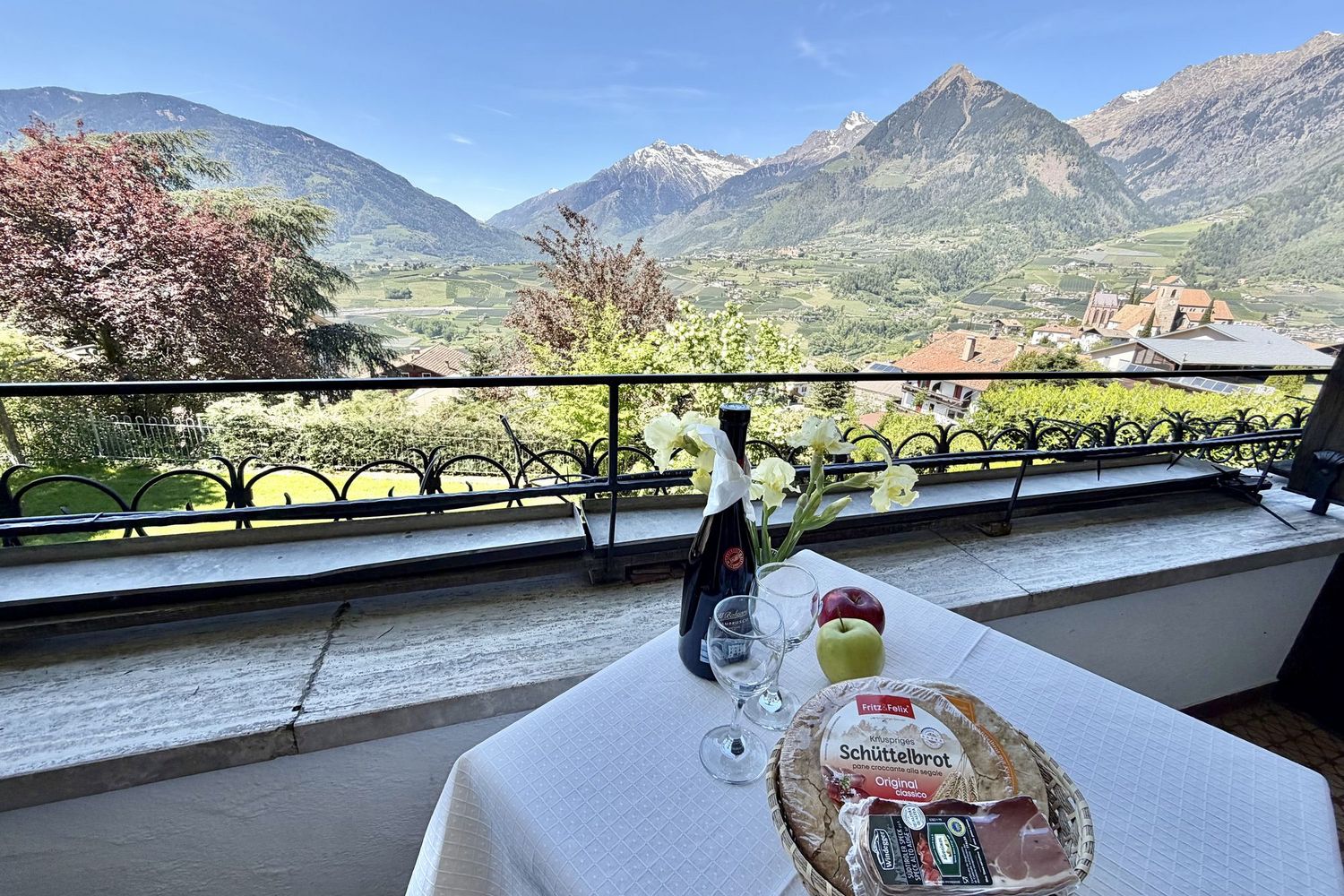 Ein Tisch auf einem Balkon mit einer Flasche Wein, zwei Gläsern, einem Korb mit Südtiroler Schüttelbrot, Speck und zwei Äpfeln vor einem Bergpanorama.