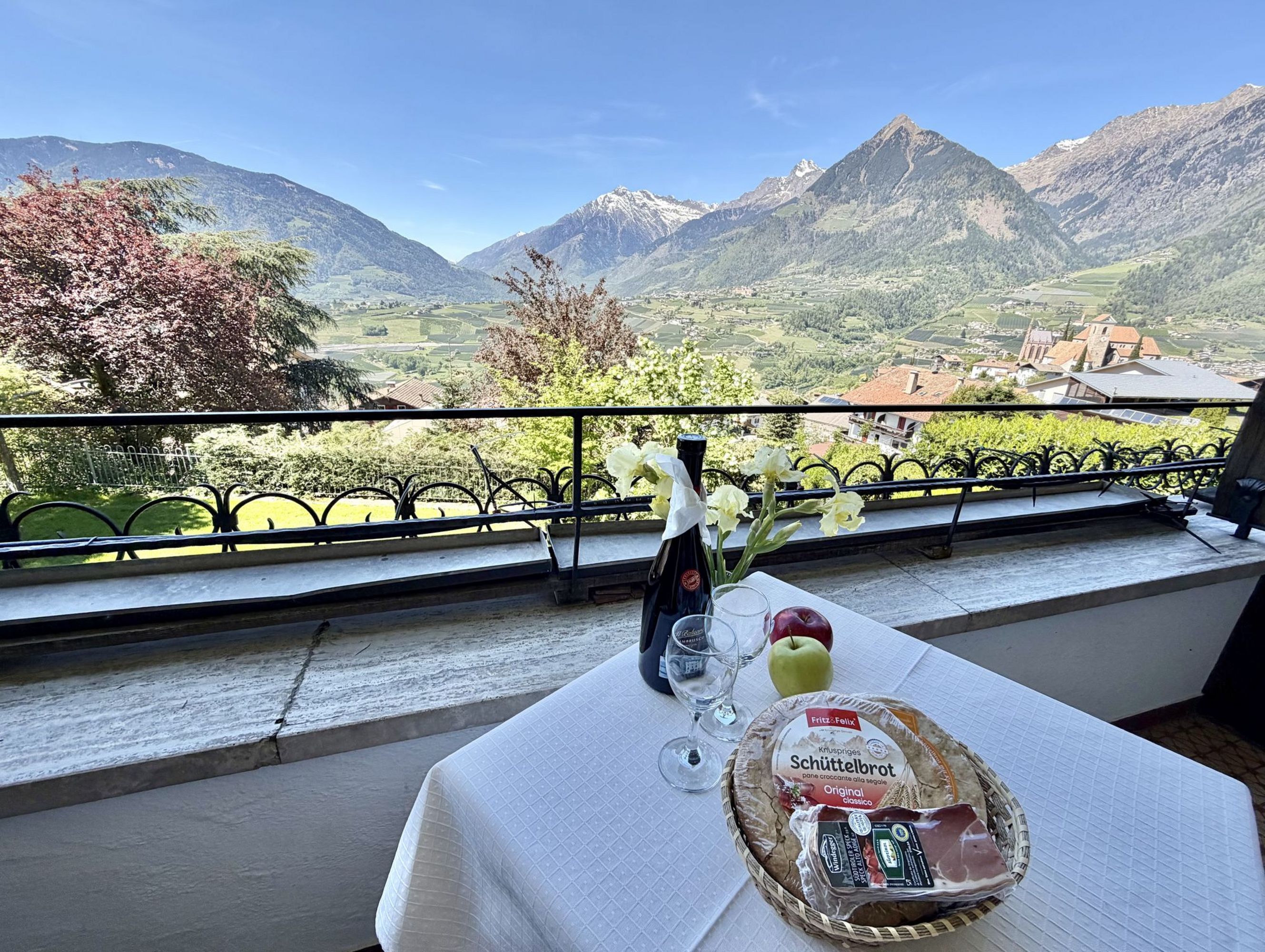 Ein Tisch auf einem Balkon mit einer Flasche Wein, zwei Gläsern, einem Korb mit Südtiroler Schüttelbrot, Speck und zwei Äpfeln vor einem Bergpanorama.