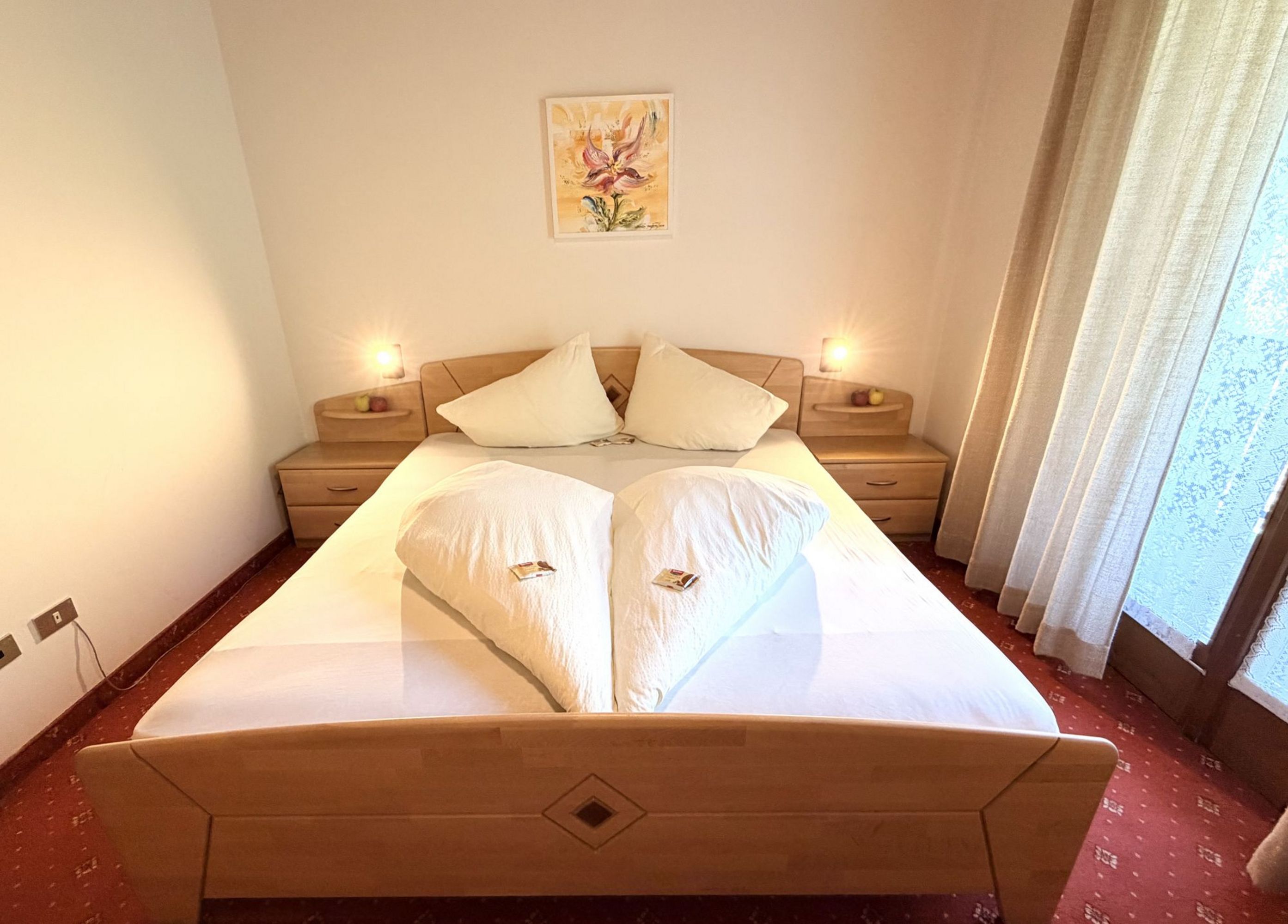 Double bed with light wooden furniture, heart-shaped pillows, and atmospheric lighting in a cozy hotel room.