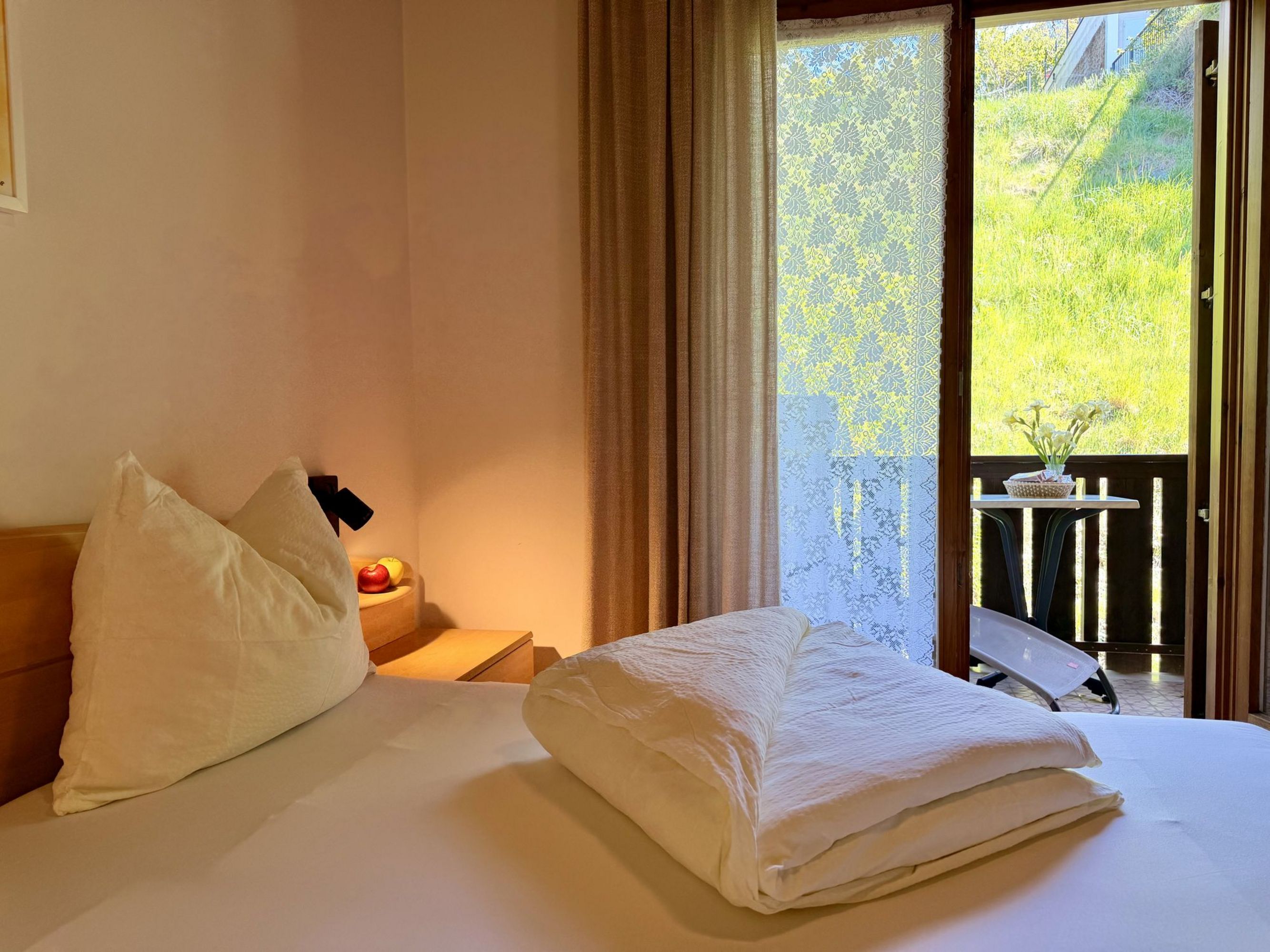 A cozy hotel room with a double bed, a nightstand with apples, and a large terrace door revealing a view of a sunny, green meadow.