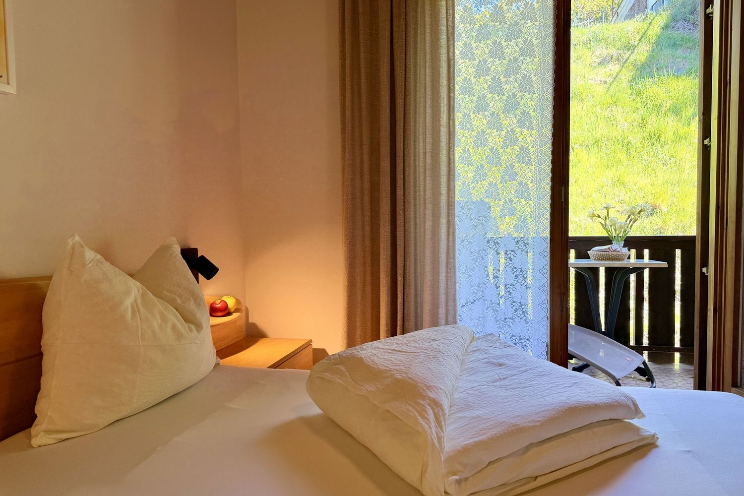 A cozy hotel room with a double bed, a nightstand with apples, and a large terrace door revealing a view of a sunny, green meadow.