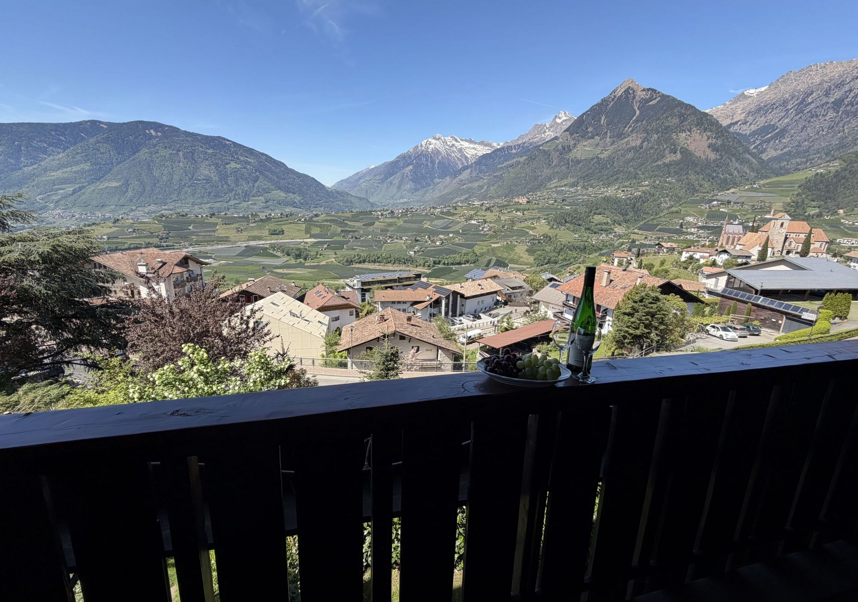 Ein Blick von einem Balkon auf eine alpine Landschaft mit Dorf, Feldern und Bergen, im Vordergrund stehen eine Weinflasche, zwei Weingläser und eine Schale mit Weintrauben.