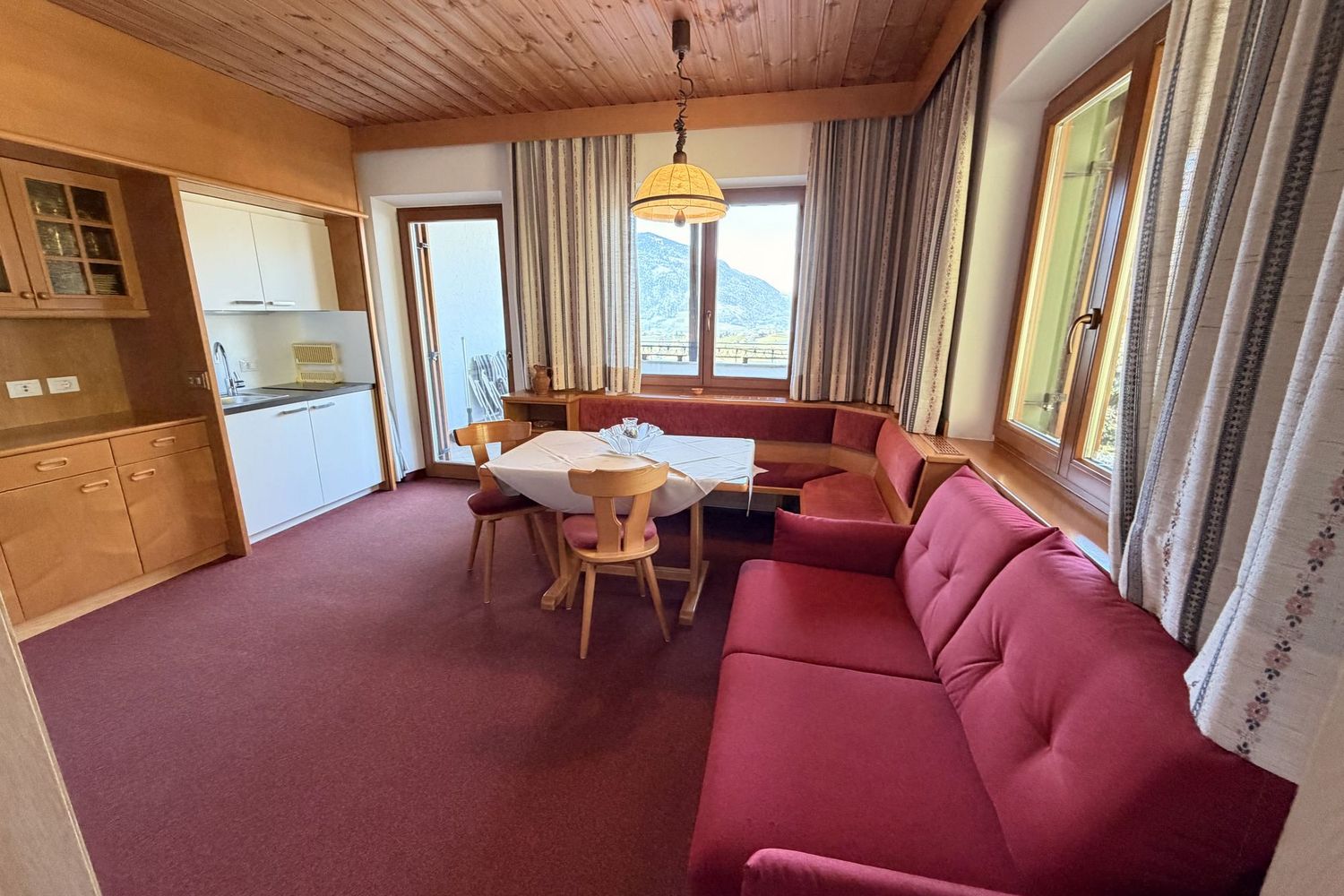 Ein gemütlicher Wohnraum mit Holzdecke, rotem Teppichboden, einer Essecke mit Eckbank und einem roten Sofa sowie einer integrierten kleinen Küchenzeile.