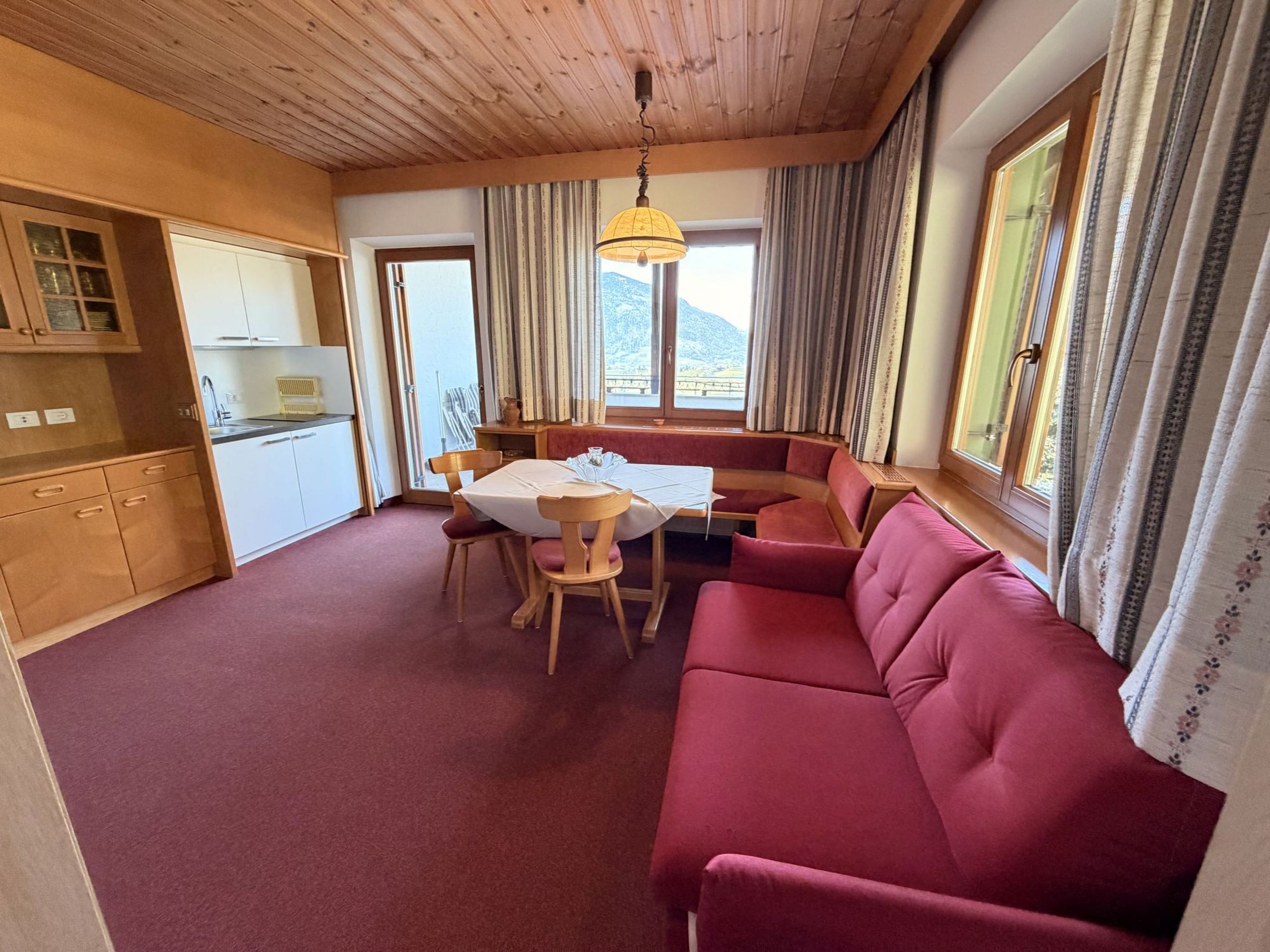 Ein gemütlicher Wohnraum mit Holzdecke, rotem Teppichboden, einer Essecke mit Eckbank und einem roten Sofa sowie einer integrierten kleinen Küchenzeile.