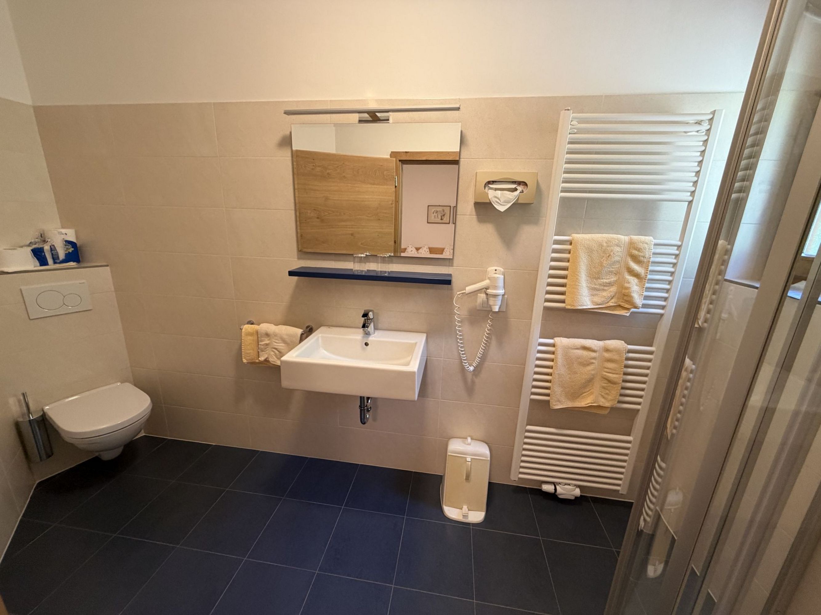 Ein modernes Badezimmer mit blauem Fliesenboden, beigen Wandfliesen, einer wandhängenden Toilette, einem Waschbecken mit Spiegel und einem Handtuchheizkörper mit darauf hängenden gelben Handtüchern.