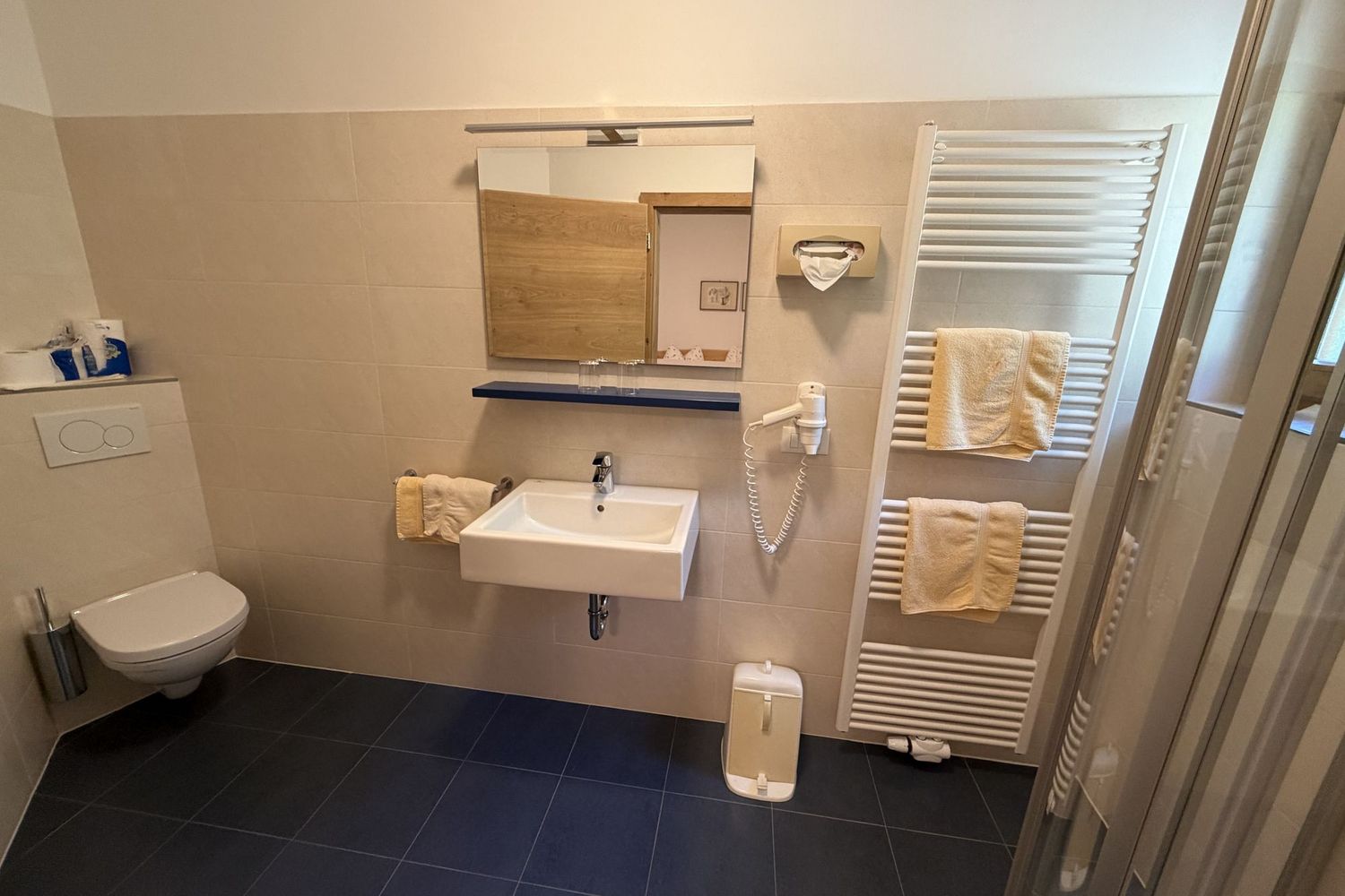 Ein modernes Badezimmer mit blauem Fliesenboden, beigen Wandfliesen, einer wandhängenden Toilette, einem Waschbecken mit Spiegel und einem Handtuchheizkörper mit darauf hängenden gelben Handtüchern.