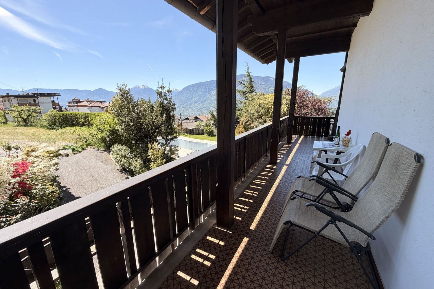 Ein überdachter Balkon mit zwei Klappstühlen, einem kleinen Tisch und einem hölzernen Geländer, mit Blick auf eine sonnige Berglandschaft.