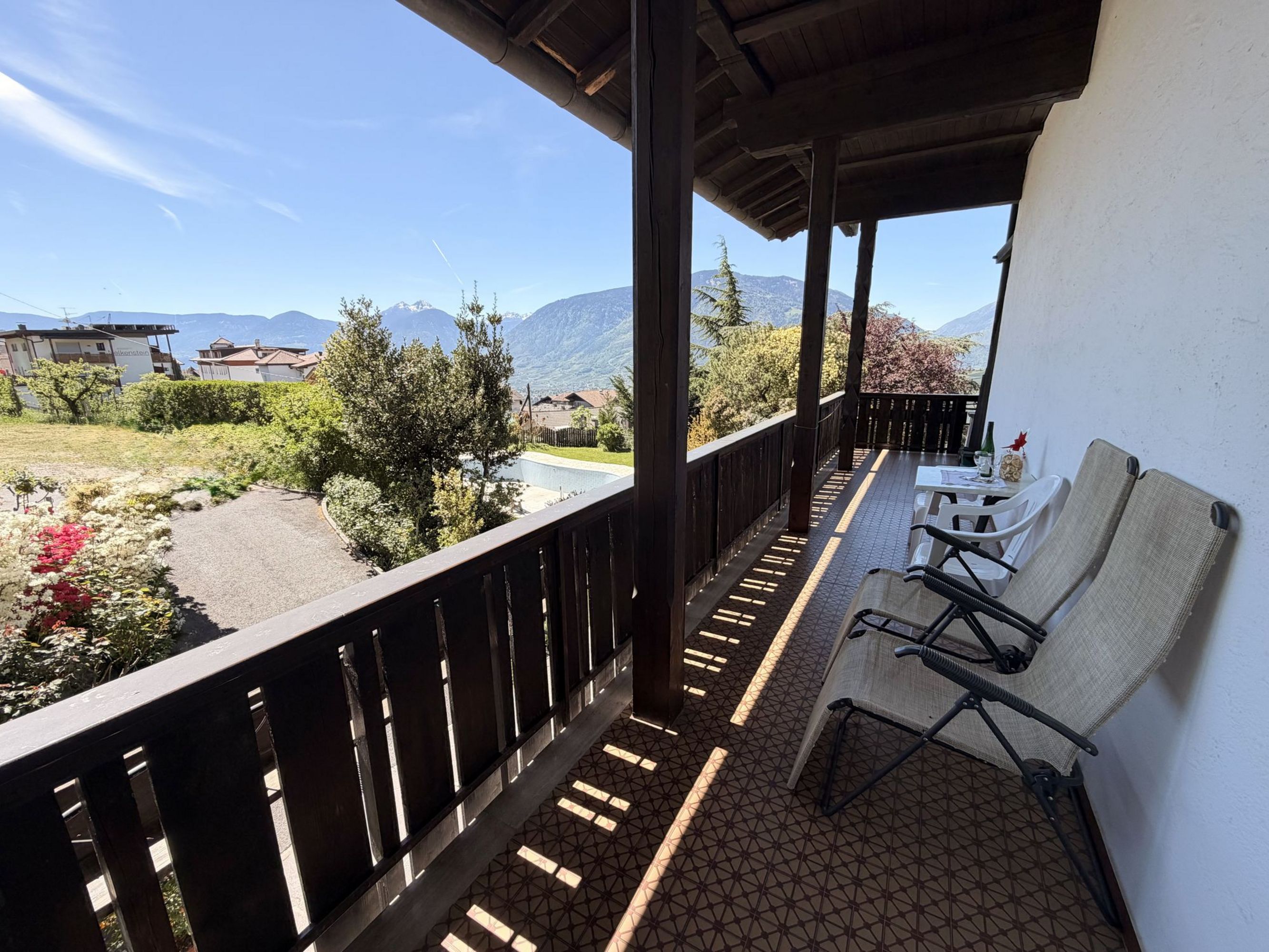 Ein überdachter Balkon mit zwei Klappstühlen, einem kleinen Tisch und einem hölzernen Geländer, mit Blick auf eine sonnige Berglandschaft.