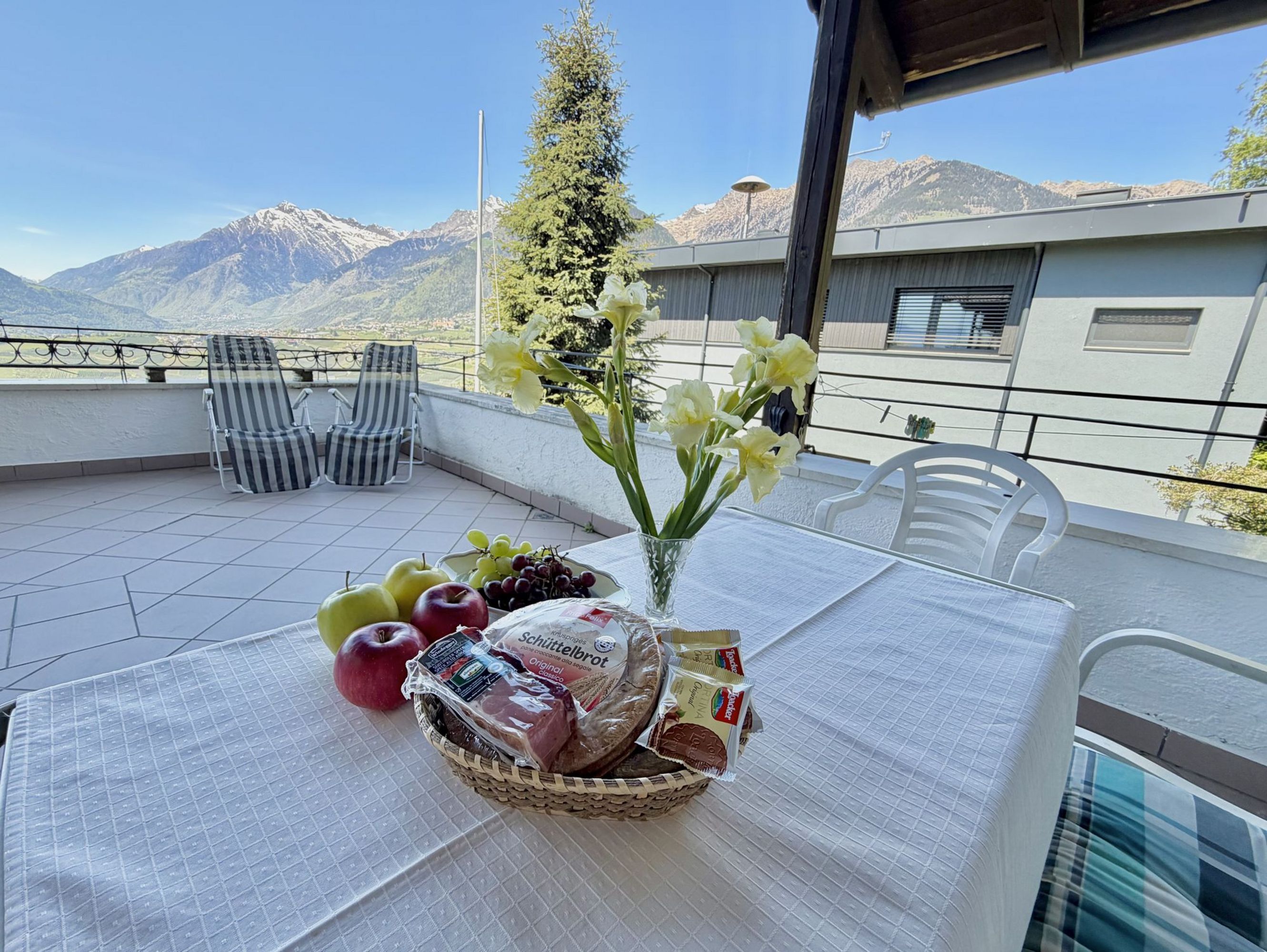 Ein weiß gedeckter Tisch auf einer sonnigen Terrasse mit Schüttelbrot, Obst und Blumen, vor einem Panorama mit Bergen und blauem Himmel.