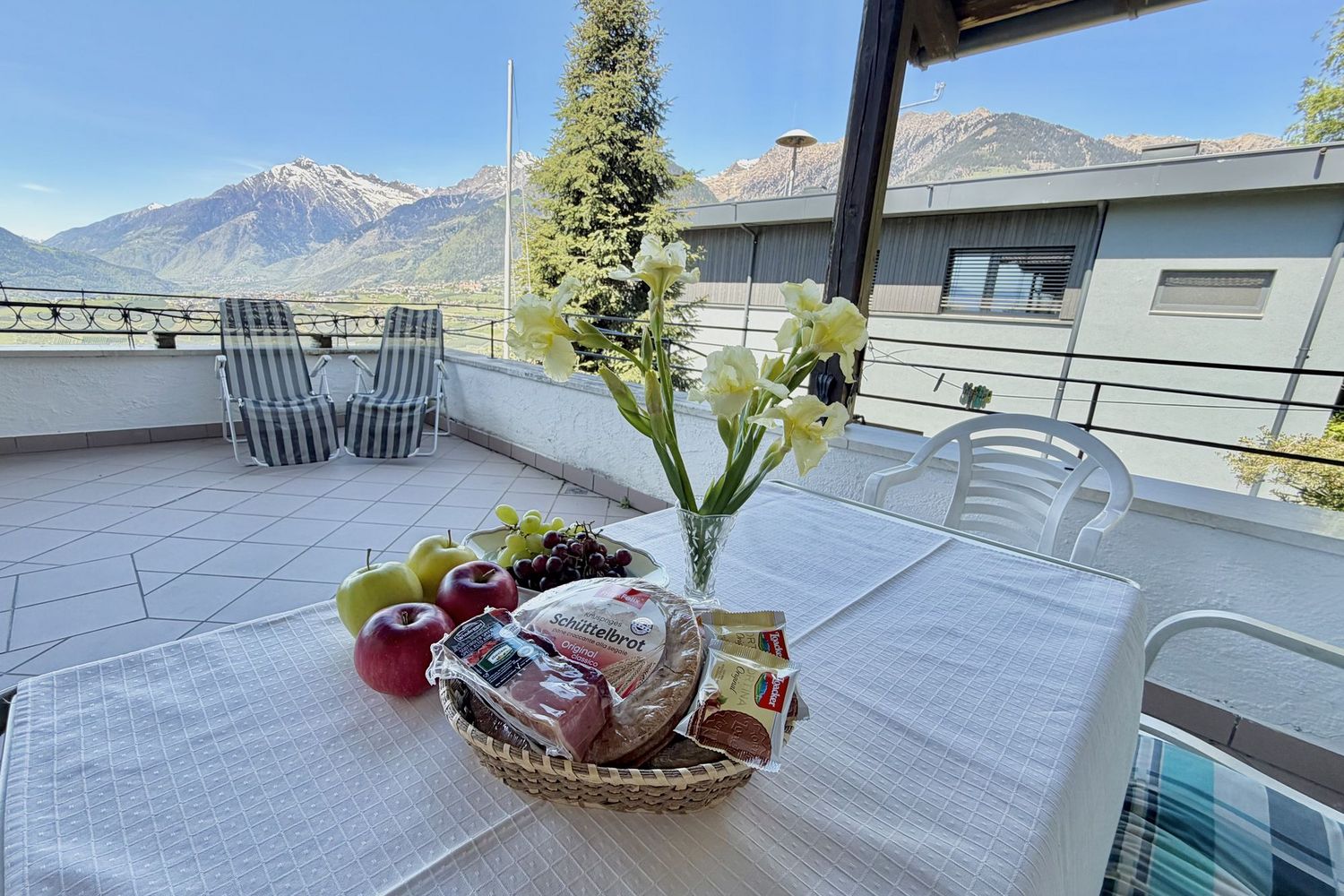 Ein weiß gedeckter Tisch auf einer sonnigen Terrasse mit Schüttelbrot, Obst und Blumen, vor einem Panorama mit Bergen und blauem Himmel.