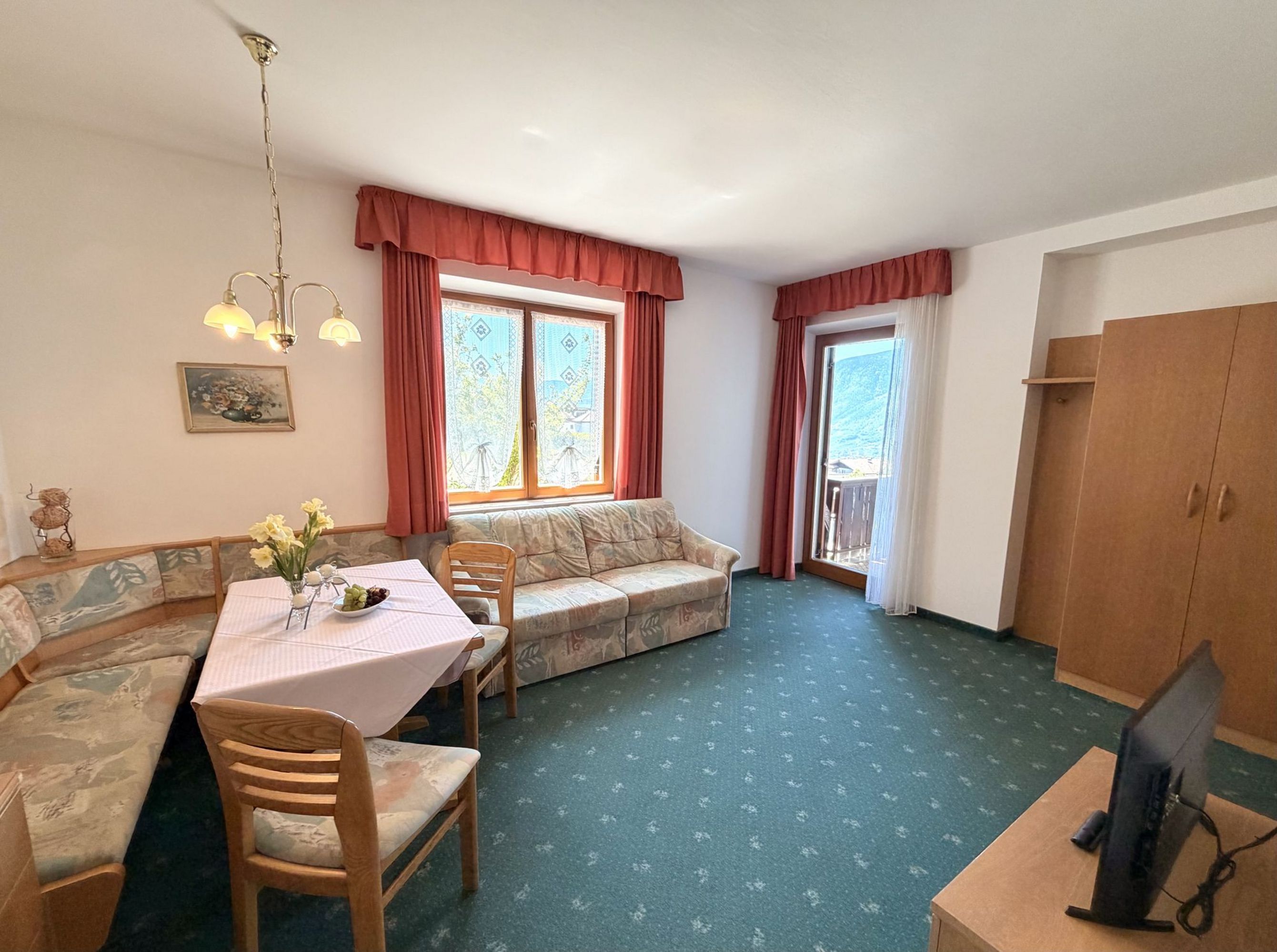 Ein gemütliches Wohnzimmer mit grüner Auslegeware, einer Eckbank, einem Sofa mit Blumenmuster, einem Esstisch und einer Terrassentür.