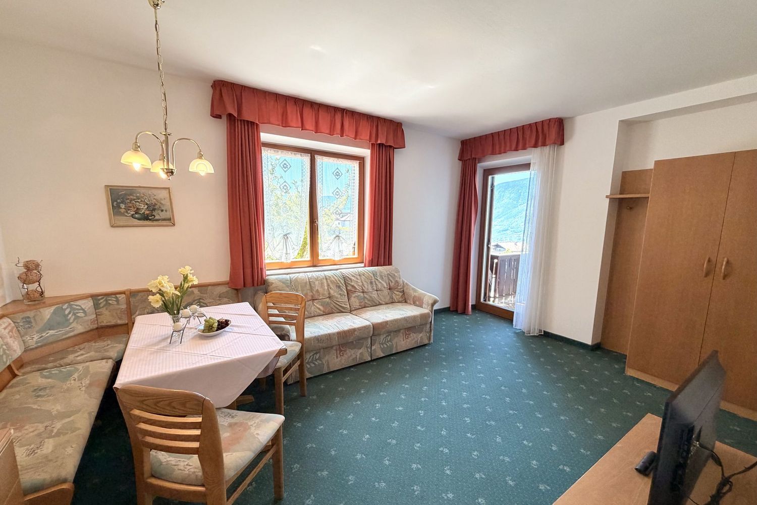 Ein gemütliches Wohnzimmer mit grüner Auslegeware, einer Eckbank, einem Sofa mit Blumenmuster, einem Esstisch und einer Terrassentür.