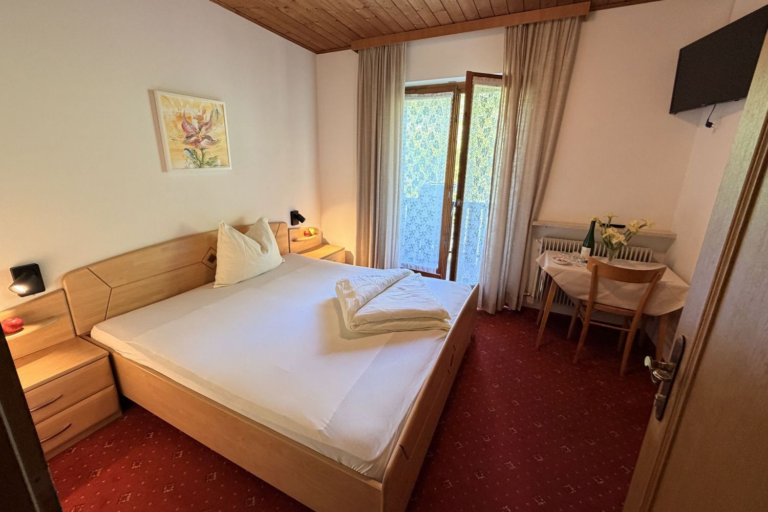 A cozy hotel room with a double bed, a small table with a chair, and balcony access on red carpeting.
