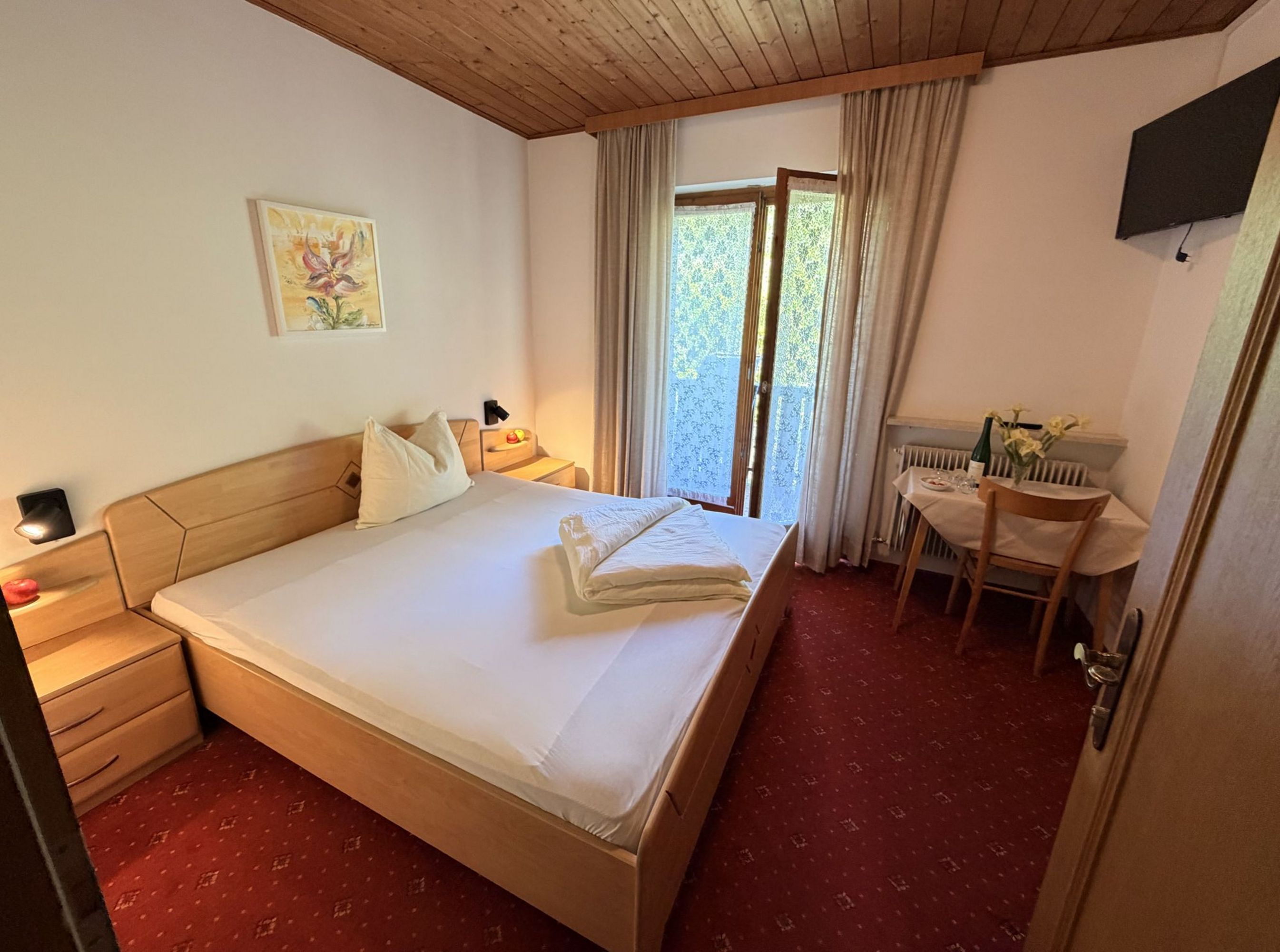 A cozy hotel room with a double bed, a small table with a chair, and balcony access on red carpeting.