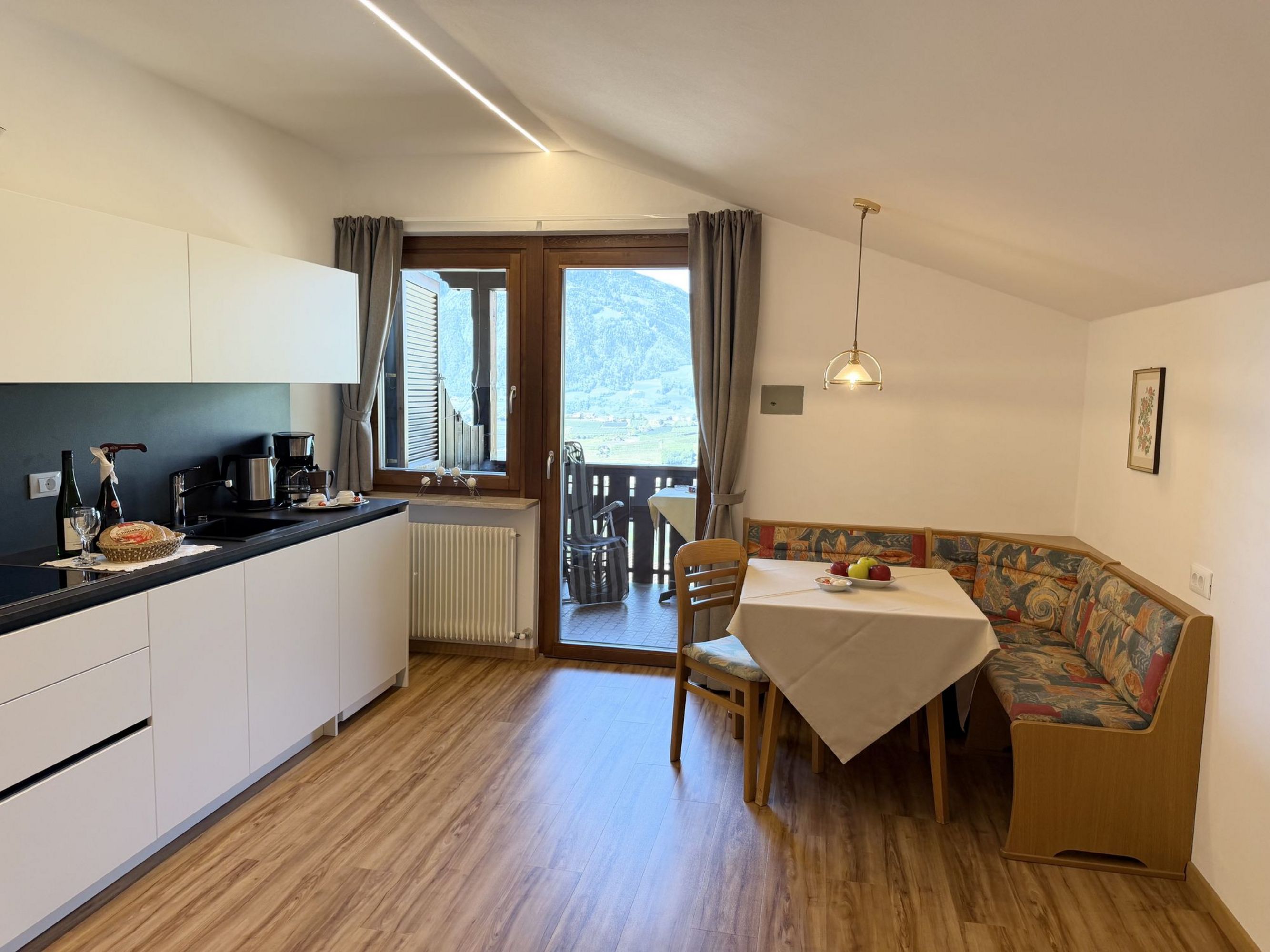 A modern eat-in kitchen with white cabinets, a wooden dining table with a corner bench, and a balcony door revealing a mountain panorama.