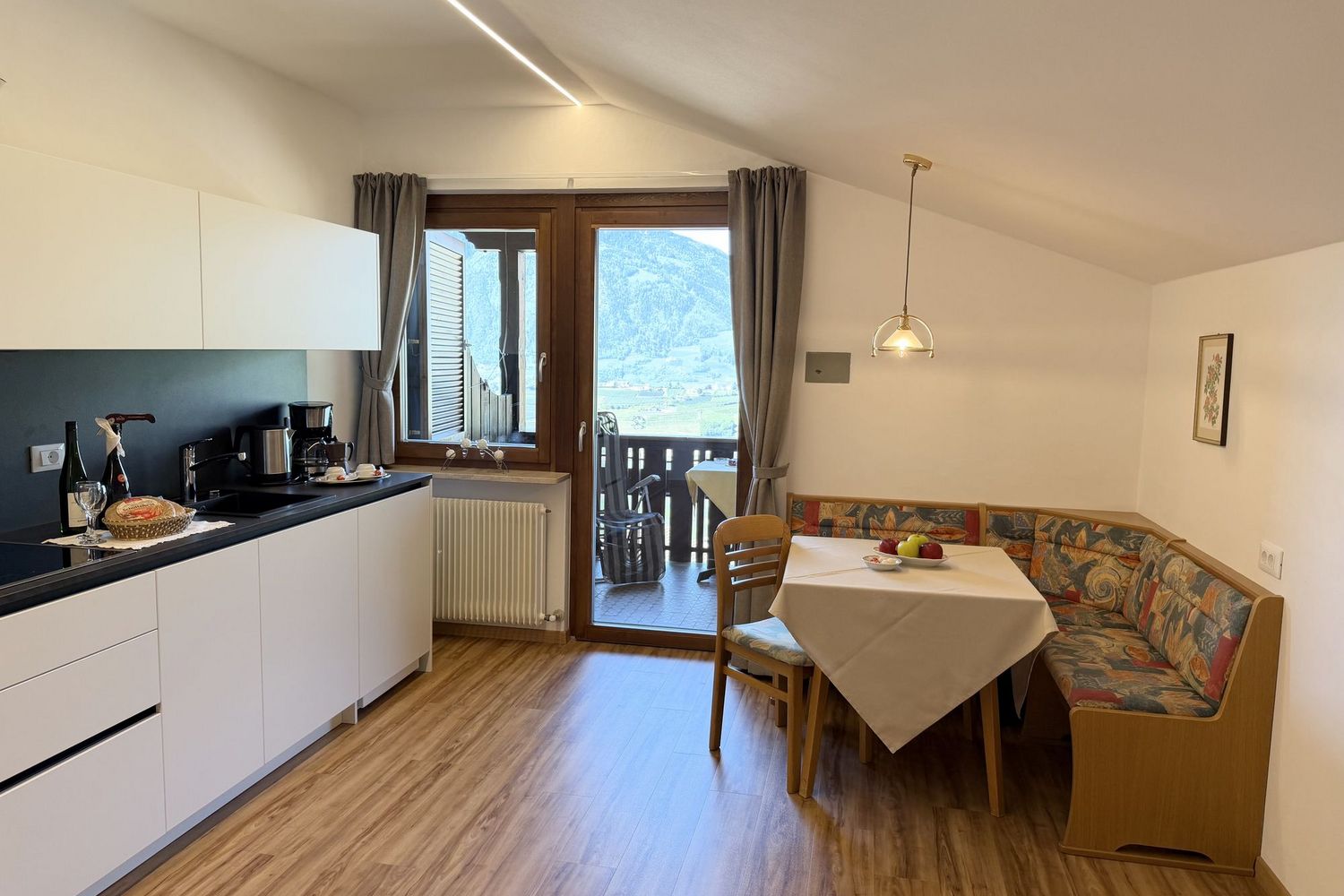 A modern eat-in kitchen with white cabinets, a wooden dining table with a corner bench, and a balcony door revealing a mountain panorama.