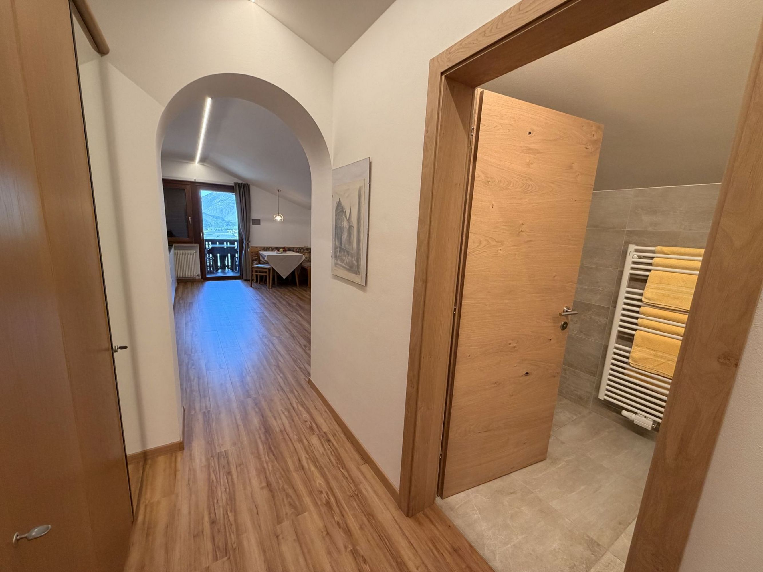 Ein Flurbereich mit hellem Holzboden, der durch einen Rundbogen in einen angrenzenden Essbereich mit Balkonzugang führt, rechts eine geöffnete Holztür zu einem Badezimmer mit Handtuchheizkörper.
