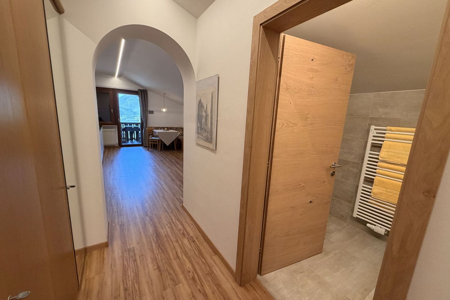 Ein Flurbereich mit hellem Holzboden, der durch einen Rundbogen in einen angrenzenden Essbereich mit Balkonzugang führt, rechts eine geöffnete Holztür zu einem Badezimmer mit Handtuchheizkörper.