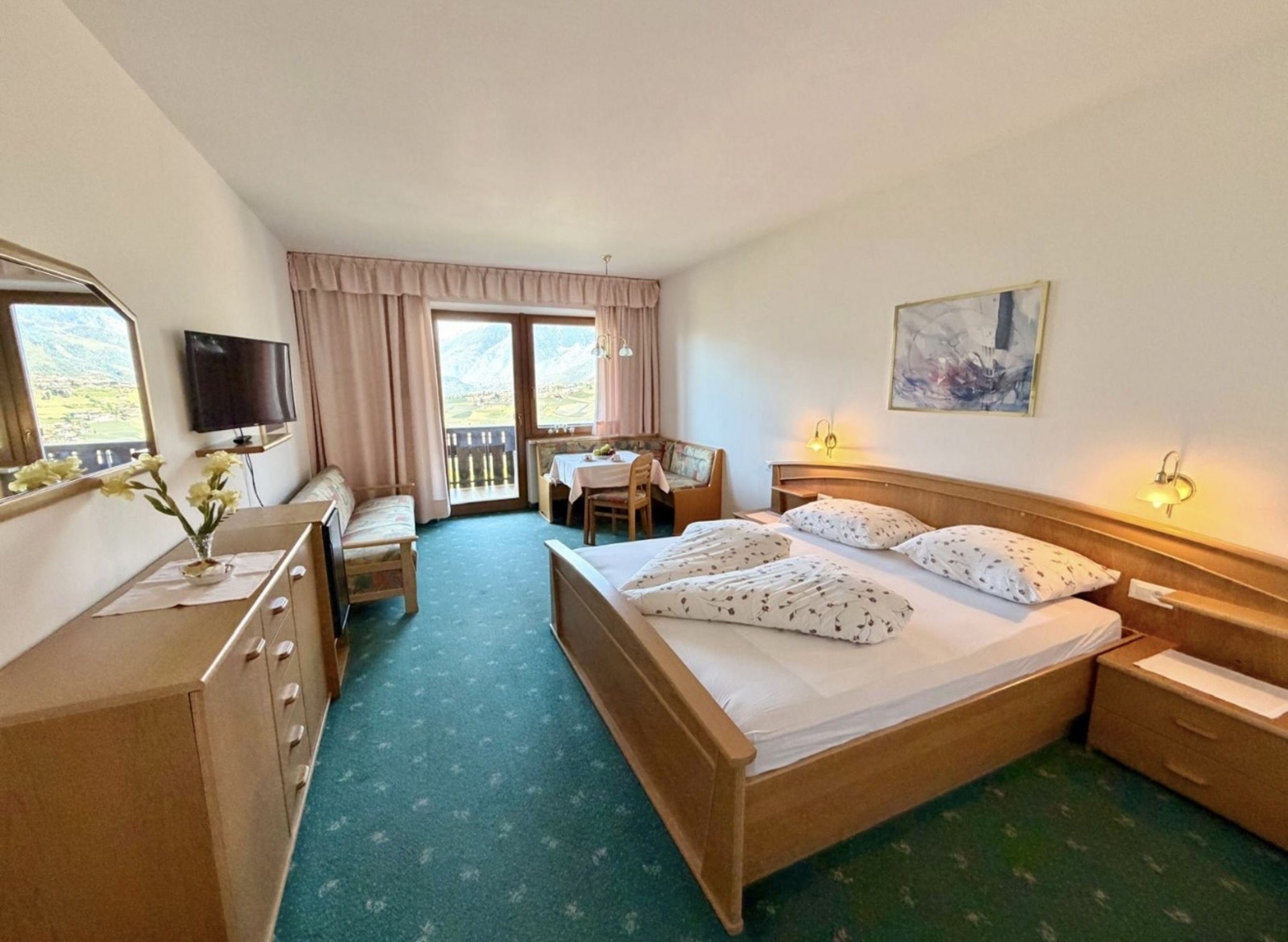 A bright hotel room with a double bed, a seating area by the window overlooking the mountains, a television, and a dresser.