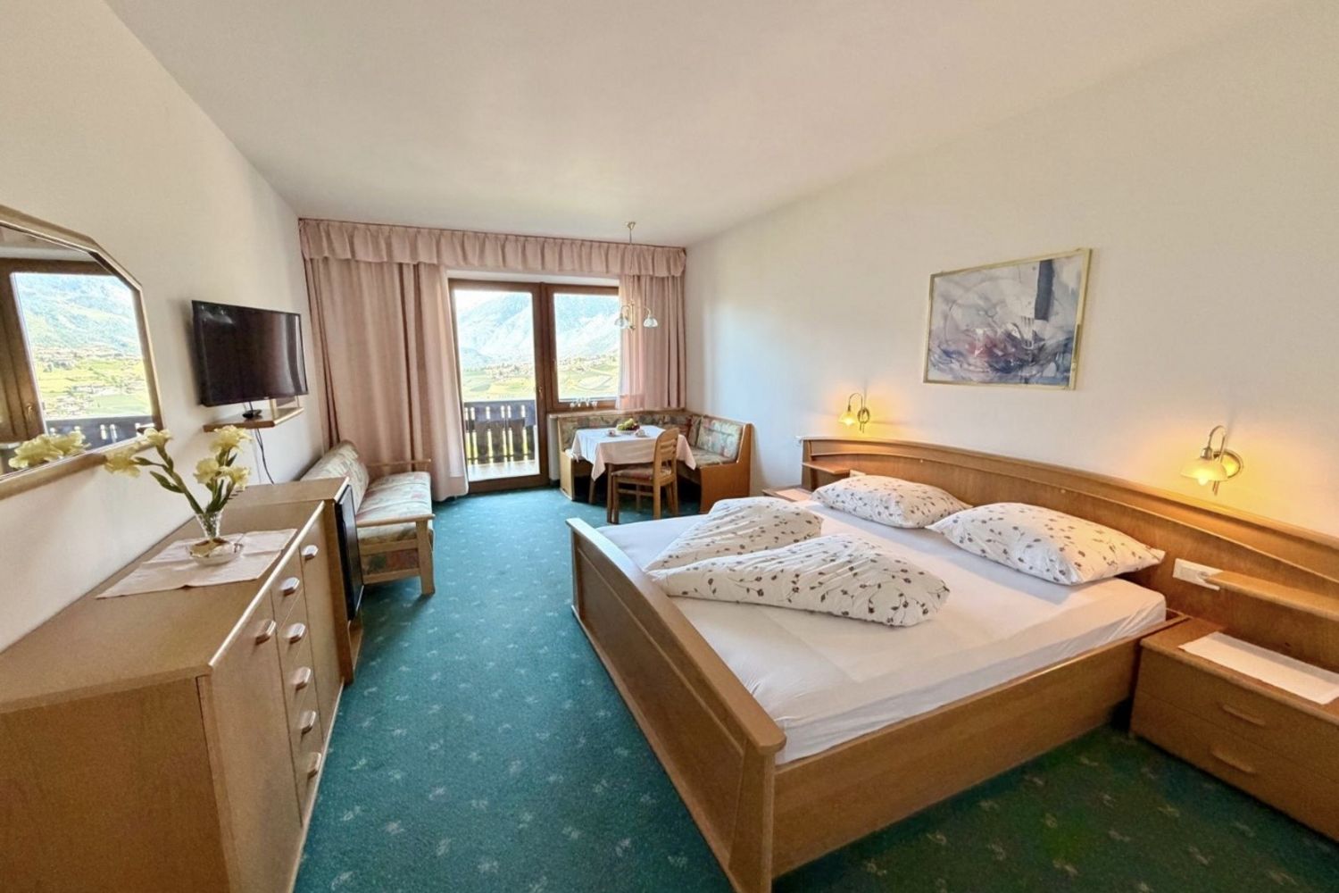 A bright hotel room with a double bed, a seating area by the window overlooking the mountains, a television, and a dresser.