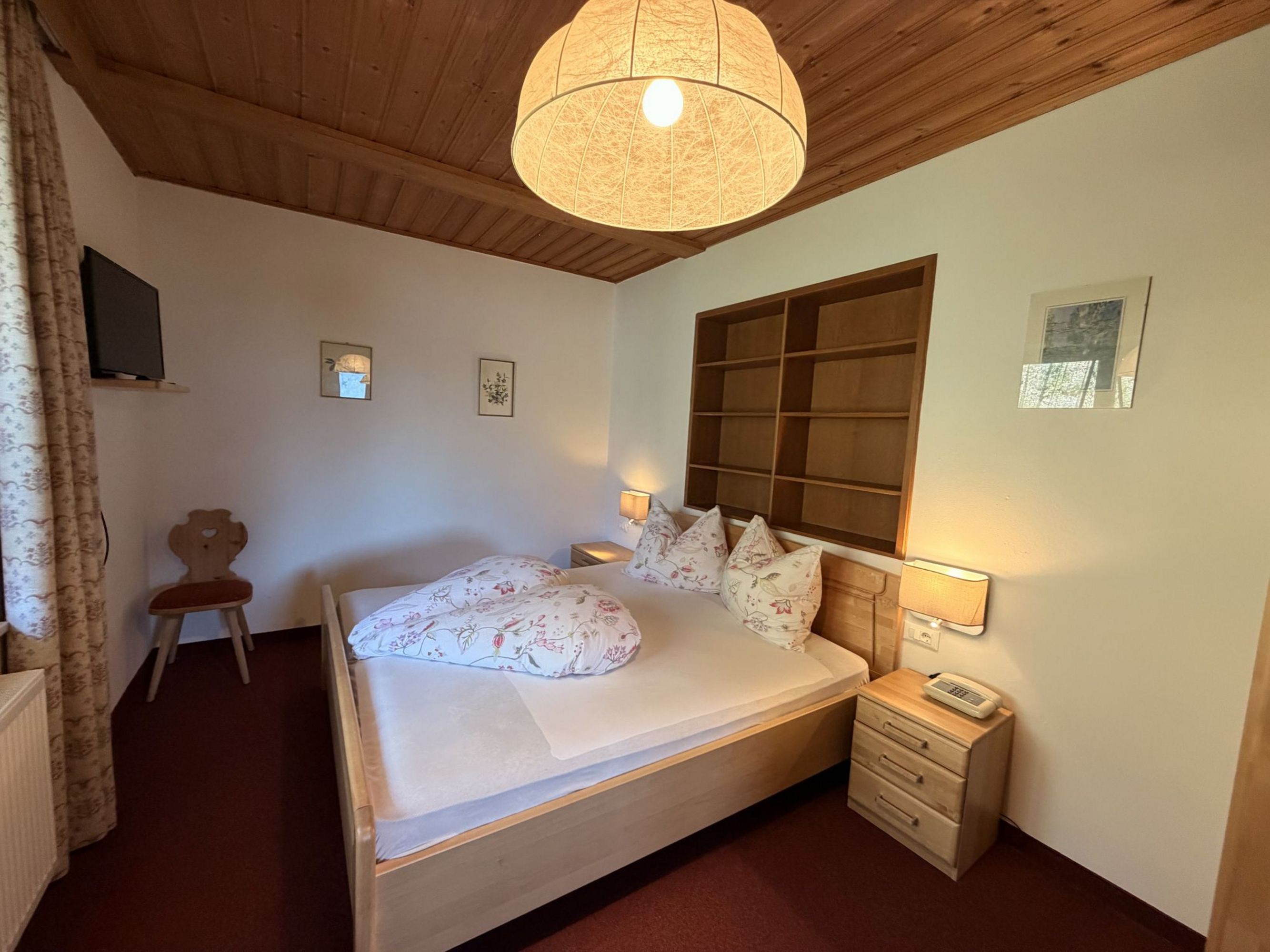 Ein gemütliches Schlafzimmer mit einem Doppelbett aus hellem Holz, zwei Nachttischlampen, einem Regal an der Wand und einer großen runden Deckenleuchte.