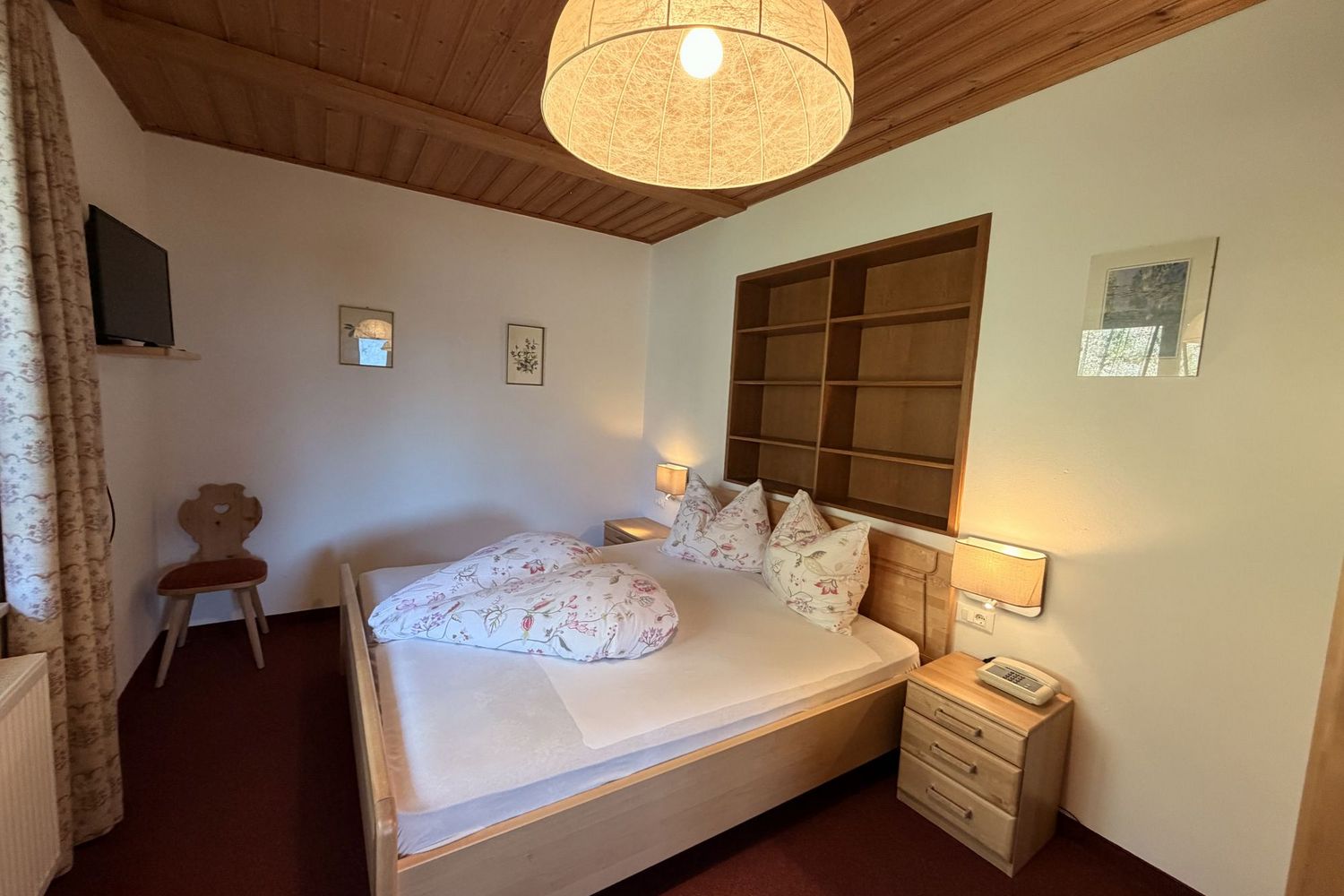 Ein gemütliches Schlafzimmer mit einem Doppelbett aus hellem Holz, zwei Nachttischlampen, einem Regal an der Wand und einer großen runden Deckenleuchte.