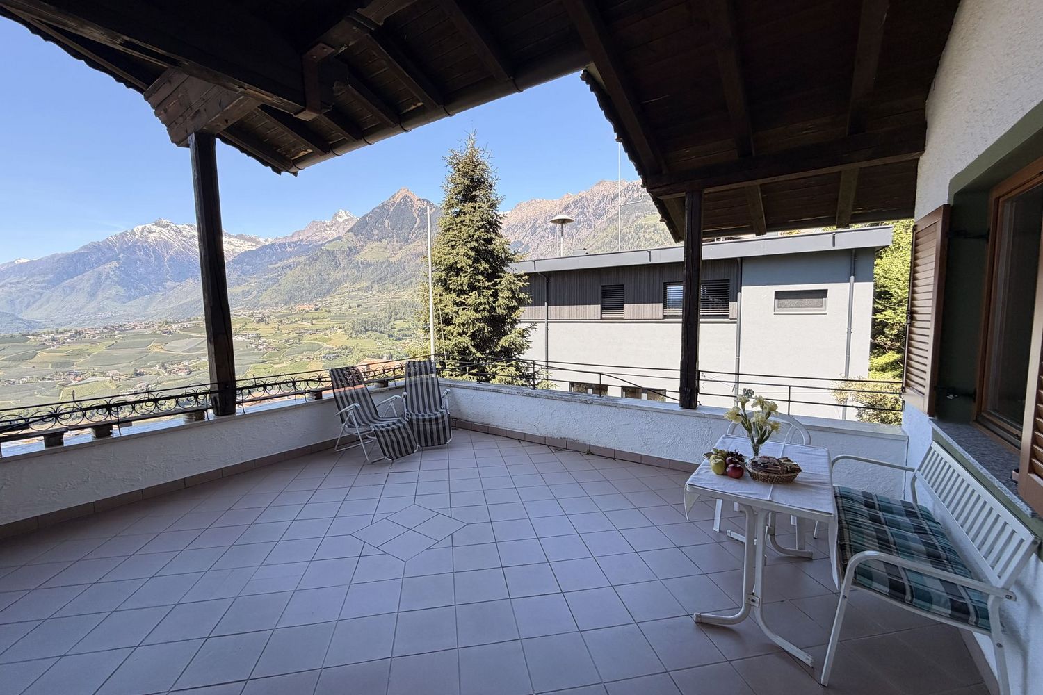 Eine überdachte Terrasse mit gefliestem Boden, einem Tisch mit Obstschale und zwei Liegestühlen vor einem Panorama aus Bergen und einem Tal.