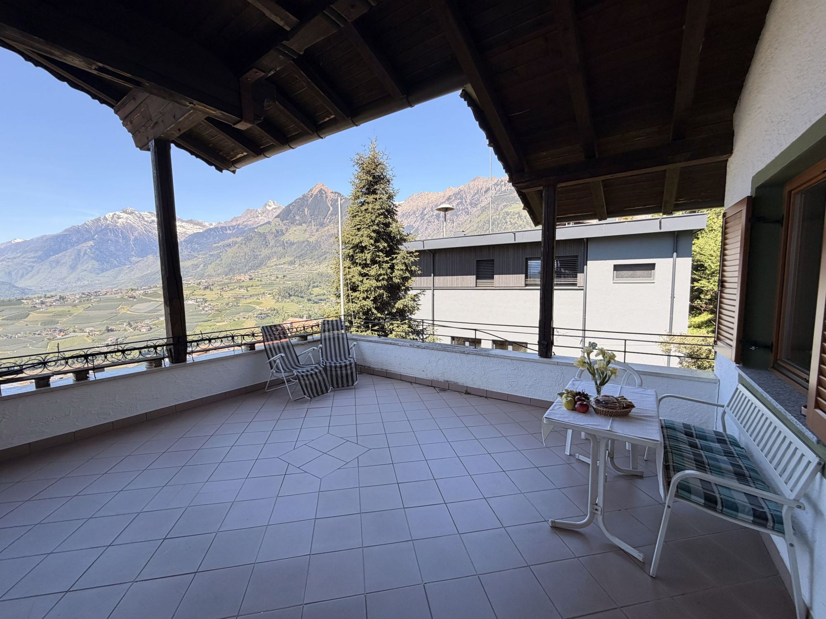 Eine überdachte Terrasse mit gefliestem Boden, einem Tisch mit Obstschale und zwei Liegestühlen vor einem Panorama aus Bergen und einem Tal.