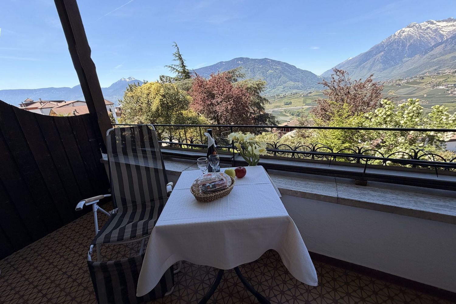 Ein kleiner Tisch mit Schüttelbrot, Obst und zwei Gläsern auf einem Balkon; im Hintergrund erstreckt sich eine sonnige Berglandschaft