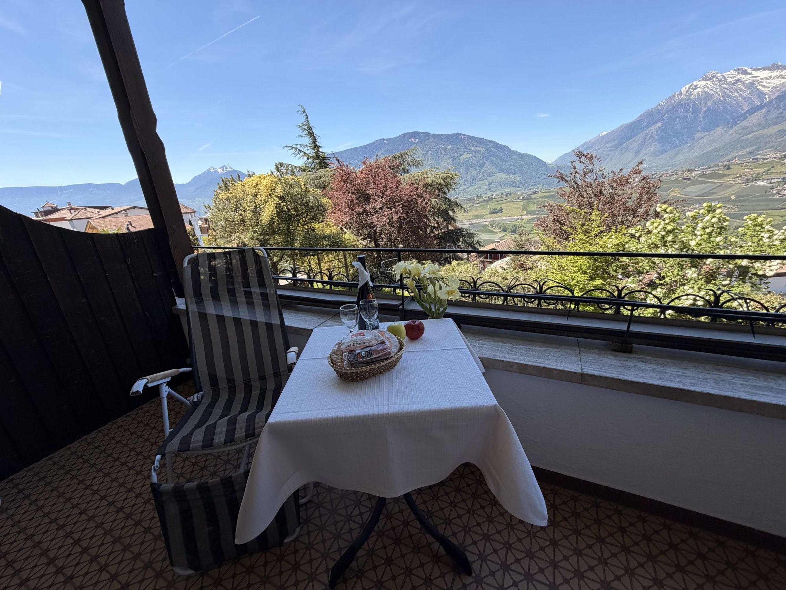 Ein kleiner Tisch mit Schüttelbrot, Obst und zwei Gläsern auf einem Balkon; im Hintergrund erstreckt sich eine sonnige Berglandschaft