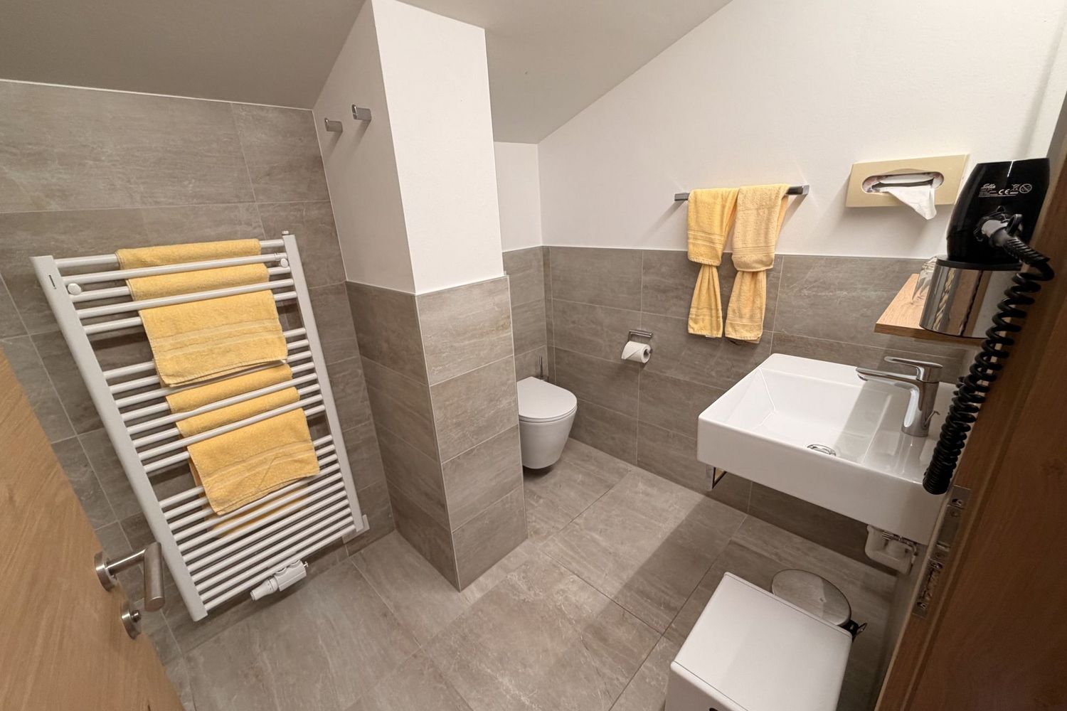 A modern bathroom with gray tiles, a white sink, a toilet, a towel radiator, and yellow towels.