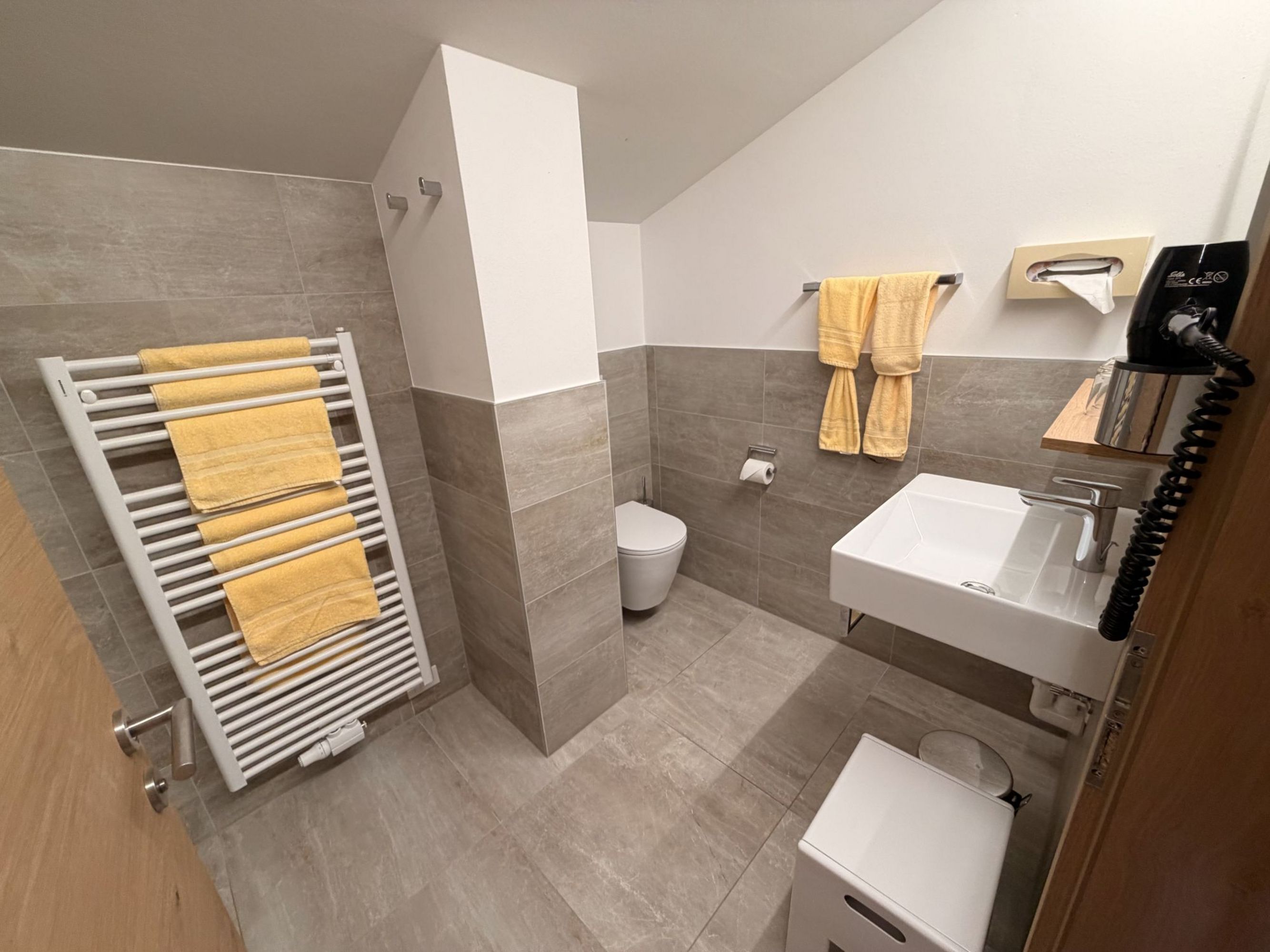 A modern bathroom with gray tiles, a white sink, a toilet, a towel radiator, and yellow towels.