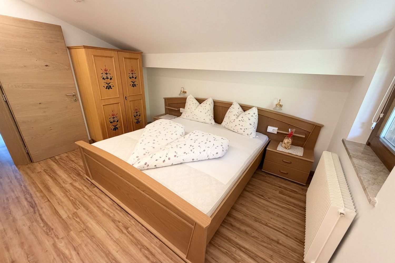 A cozy bedroom with a sloped ceiling, a rustic wooden bed with floral bedding, a matching wardrobe with floral decorations, and light wooden floors.