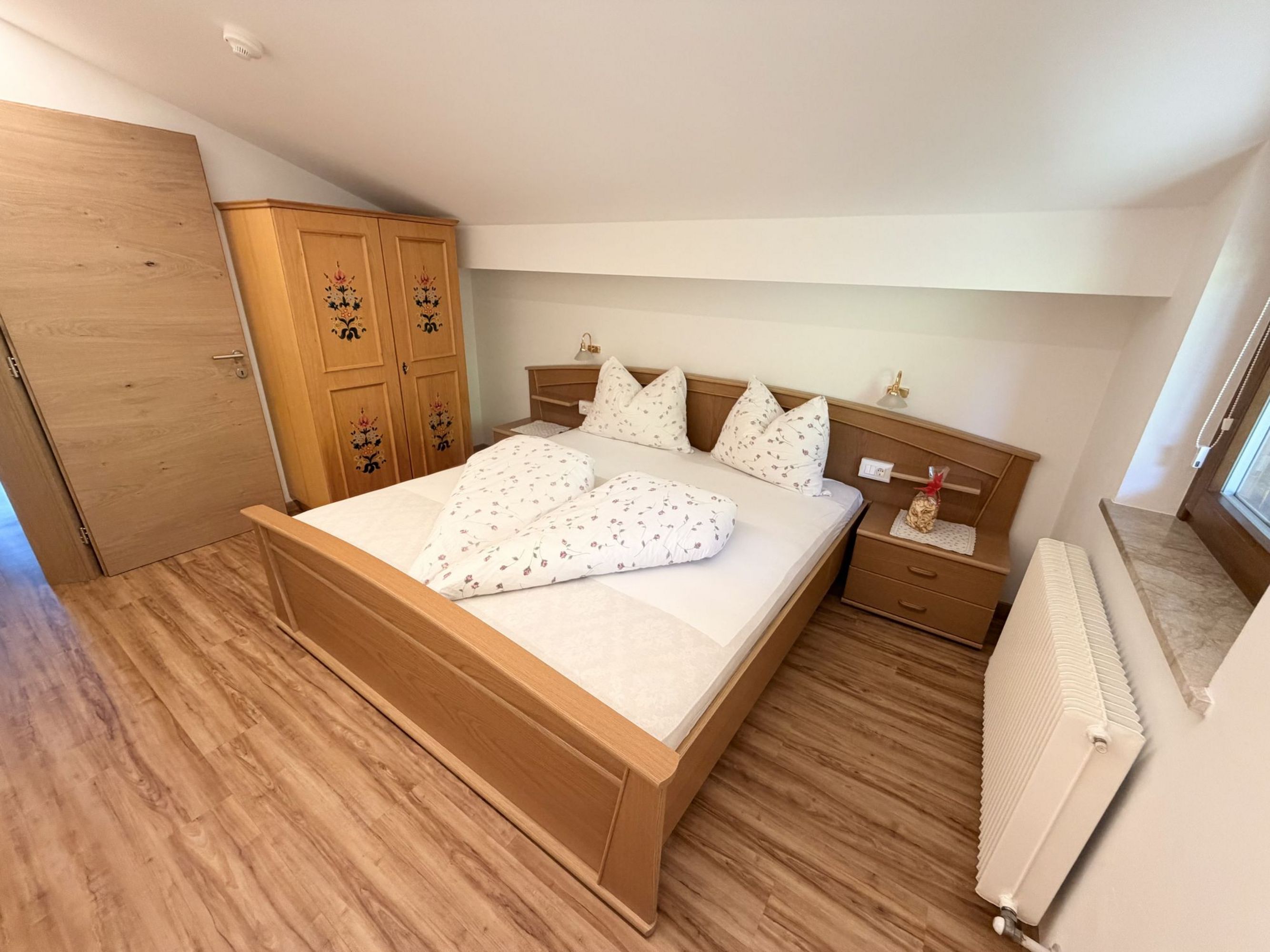 A cozy bedroom with a sloped ceiling, a rustic wooden bed with floral bedding, a matching wardrobe with floral decorations, and light wooden floors.