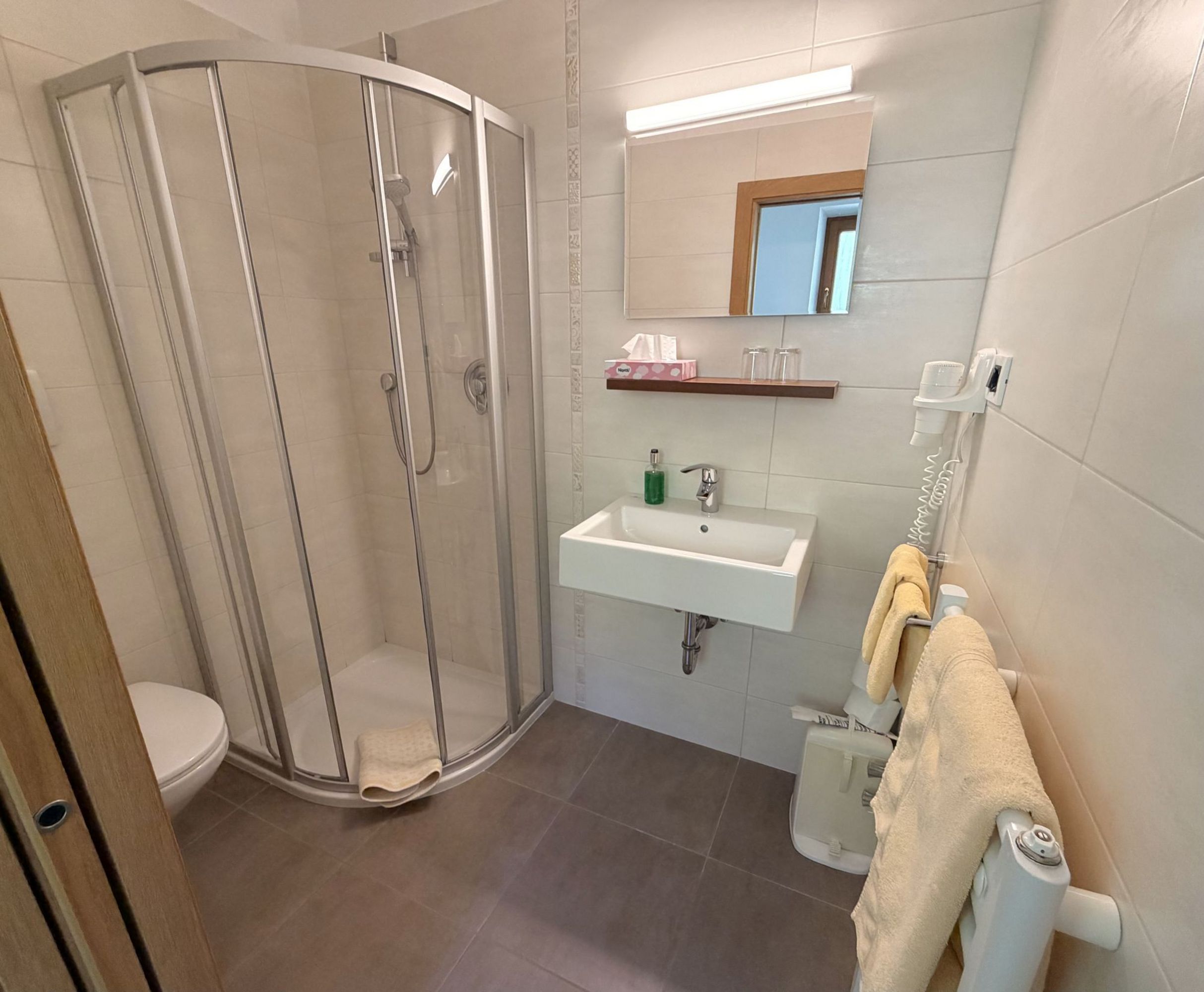 Ein modernes, hell gefliestes Badezimmer mit einer Duschkabine aus Glas, einem Waschbecken und einem Handtuchheizkörper mit hängenden Handtüchern.