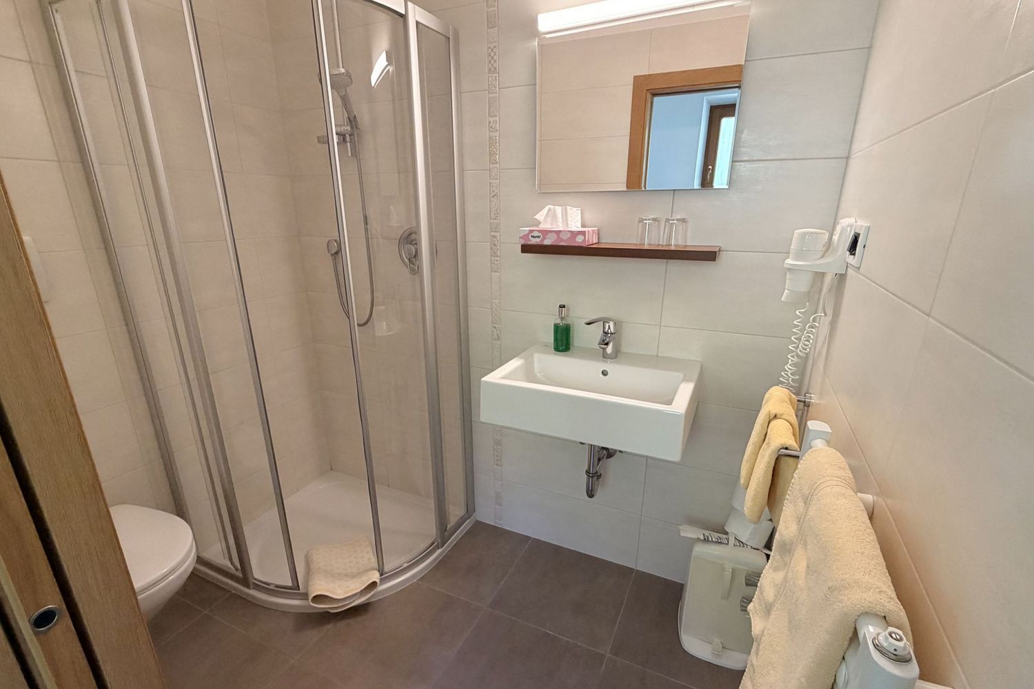 Ein modernes, hell gefliestes Badezimmer mit einer Duschkabine aus Glas, einem Waschbecken und einem Handtuchheizkörper mit hängenden Handtüchern.